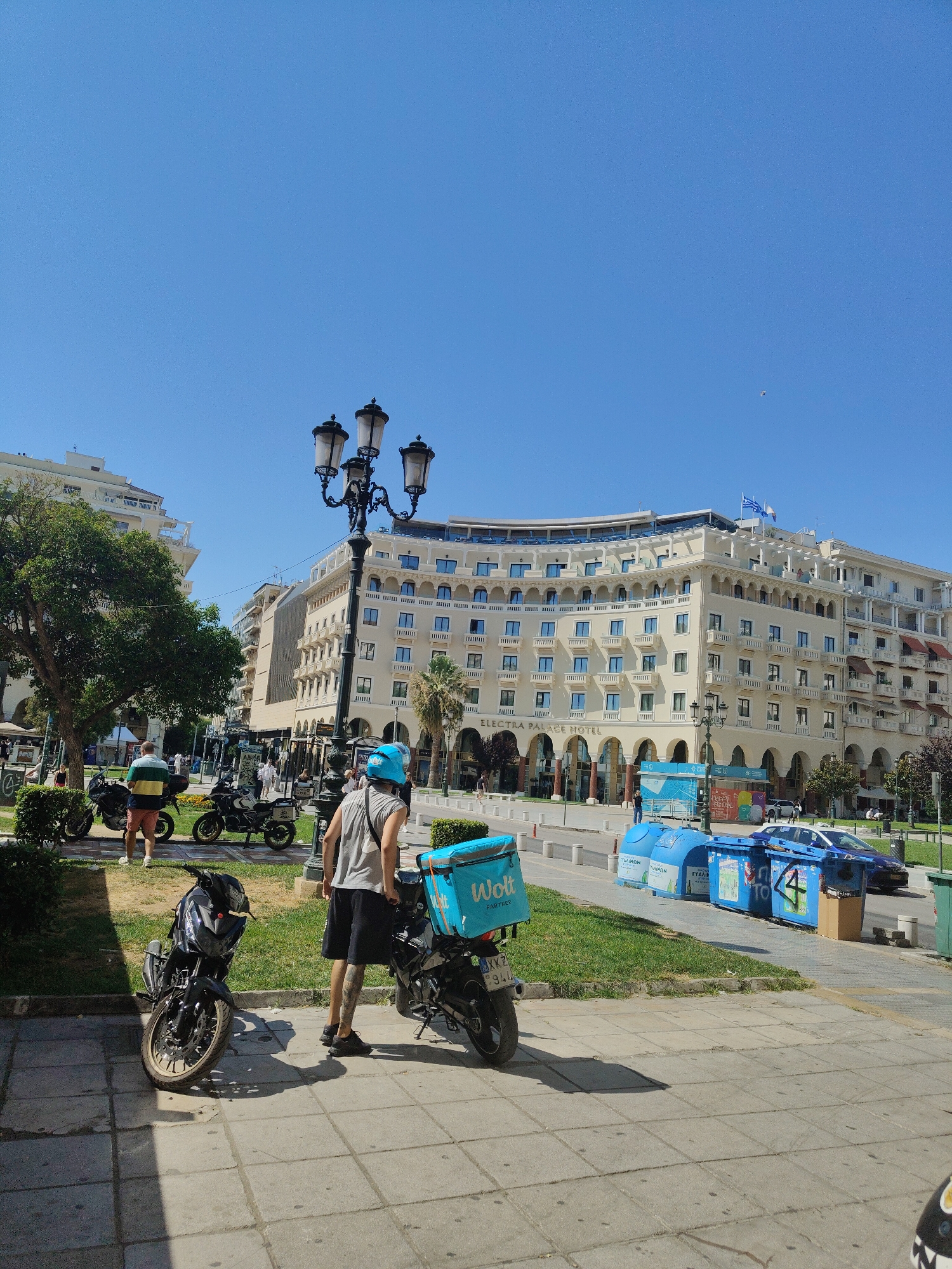 The image depicts an outdoor scene in Thessaloniki, Greece, likely a public square or sidewalk in front of the Electra Palace Hotel. A person wearing a blue helmet and a grey tank top stands next to a motorcycle with a teal delivery box labeled "Wolt Partner". Another motorcycle is parked nearby. In the background, the Electra Palace Hotel is visible, a large, light-colored building with multiple floors and arched windows. Several other people are seen walking in the background, and there are trees and streetlights. Blue recycling bins are positioned along the sidewalk. The sky is clear and blue.