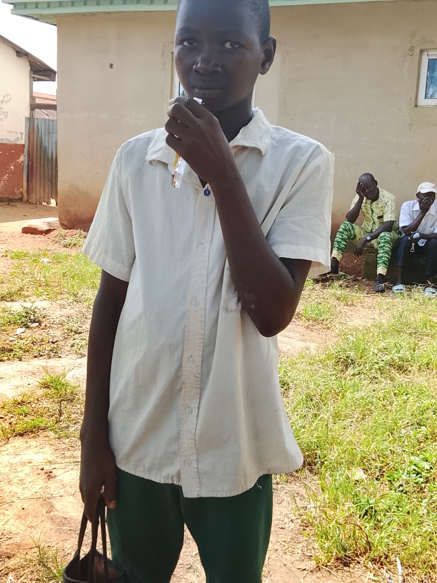 In Akwanga, Nigeria, a young person stands outdoors, holding a small, colorful object to their mouth with their right hand. They are wearing a white short-sleeved collared shirt and dark green trousers. Their left hand is holding the handle of a dark bag. In the background, two adult men are seated. The man on the left wears a patterned yellow and green shirt and green and white patterned trousers. The man on the right wears a white shirt and a white cap. The environment appears to be a dusty, outdoor area with sparse grass and a building in the background.