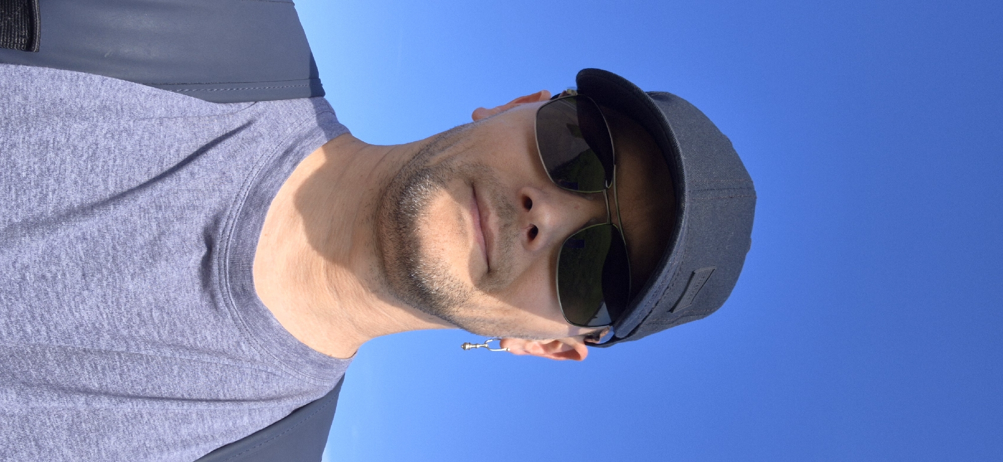 The image shows a close-up, selfie-style shot of a person wearing a black baseball cap and dark sunglasses, against a clear blue sky. The person has a light beard and is wearing a grey t-shirt. A silver earring is visible on their right ear. A portion of a dark backpack is visible on their left shoulder. The lighting suggests it is daytime. There are no discernible landmarks or features that would indicate a specific location within Gdańsk, Poland.
