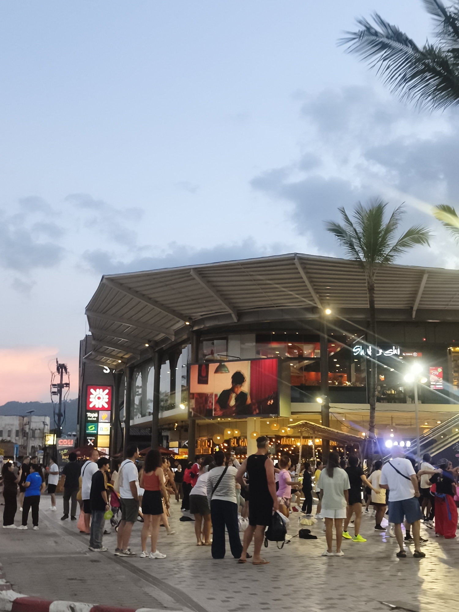 The image depicts a lively street scene in the city of Ko Samui District, Thailand, likely during the evening. A group of people are gathered on a paved walkway in front of a large, modern building with a distinctive roof structure. The building appears to be a shopping center or complex, housing various businesses with illuminated signage, including "Tops Food Hall" and what seems to be a sushi restaurant. Palm trees are visible, adding to the tropical ambiance, and the sky shows the soft colors of twilight with some clouds.
The people in the image are casually dressed, suggesting a relaxed atmosphere. They are engaged in various activities: some are walking, others are standing and talking, and a few are participating in what looks like a group exercise class, with individuals in coordinated poses. The lighting in the scene is a mix of natural twilight and artificial lights from the building and street lamps, creating a warm and inviting glow. The foreground shows a distinct red and white patterned curb, indicating the edge of the walkway. The overall impression is one of a vibrant urban hub where locals and possibly tourists are enjoying the evening.
