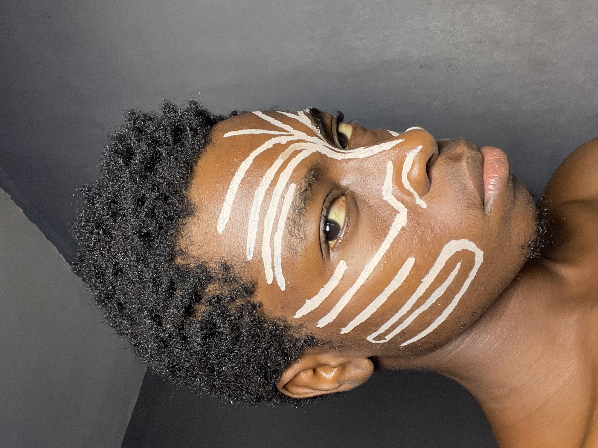 A close-up, head and shoulder shot shows a young Black person with dark, curly hair, adorned with white face paint in a tribal or artistic pattern. The paint consists of thick, white stripes across the forehead, cheeks, and extending towards the nose. The subject's gaze is directed slightly away from the camera, with their right eye more visible. The background is a plain, grey wall, suggesting an indoor setting. There is no indication of the specific location within Nkpor, Nigeria. The lighting is directional, casting shadows and highlighting the contours of the face.