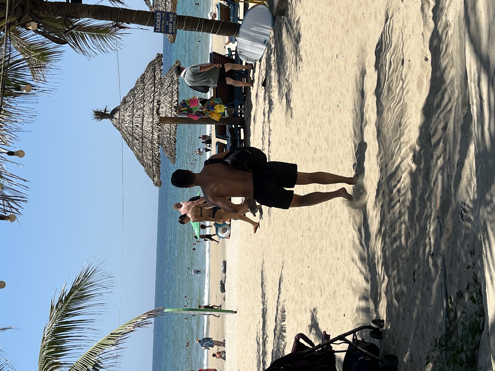 The image depicts a beach scene in Đà Nẵng, Vietnam, characterized by light-colored sand, a clear blue sky, and a calm sea. People are observed walking, swimming, and relaxing on the beach. There is a thatched-roof structure providing shade, with a locker sign visible nearby. Palm trees line the beach, creating shadows on the sand. A stroller is seen in the foreground.