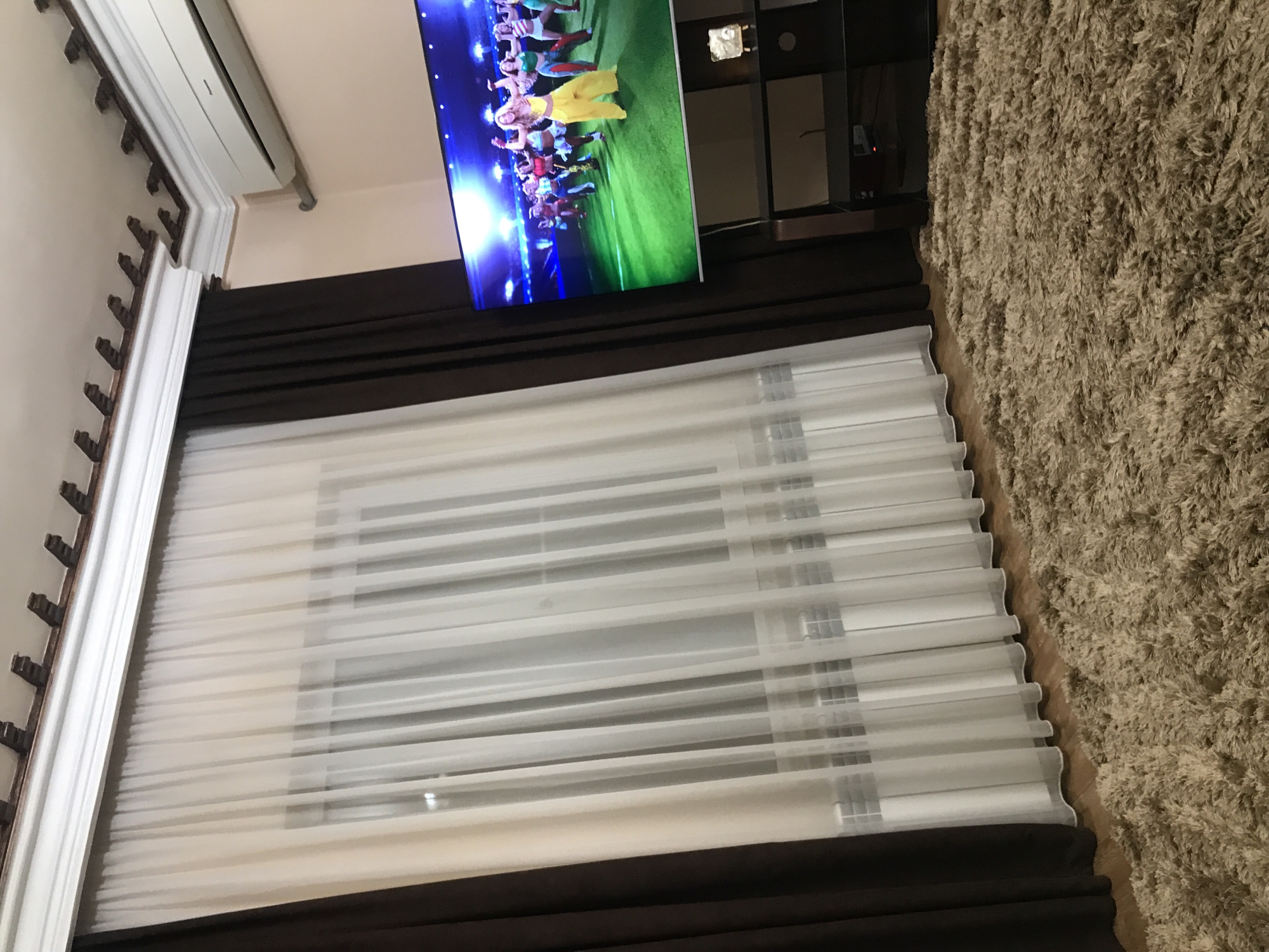 The image shows an interior view. A television is mounted on the wall above window blinds with brown valances. The television displays a video of people dancing. A shag rug lies on the floor. The location is unknown and cannot be determined from the image. The image provides no information about the people, subjects, objects, actions, interactions, or the environment that would permit identification of its location in Yerevan, Armenia.