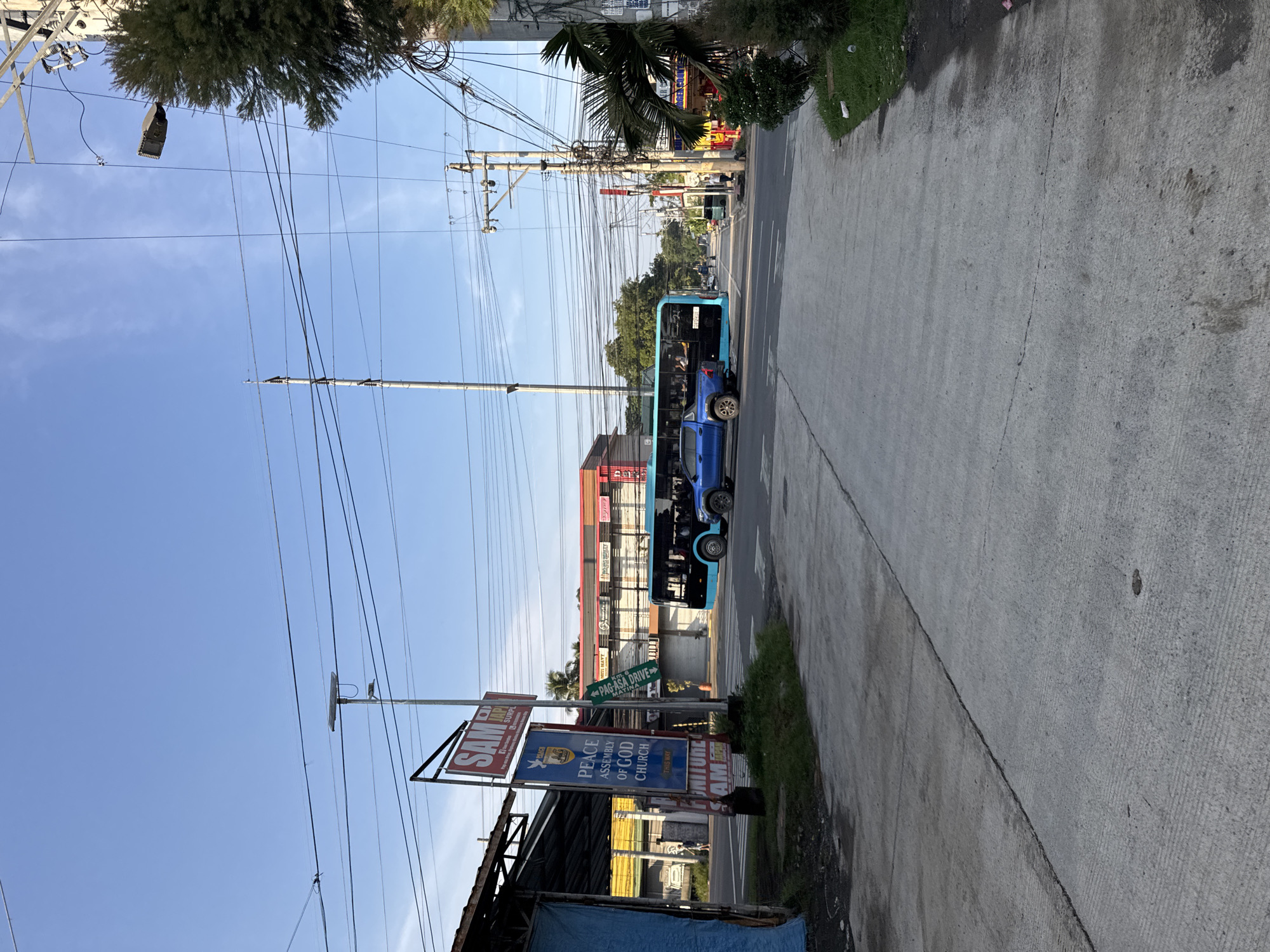 This image captures a bright, sunny daytime scene along a street in Davao City, Philippines. The main subject is a blue bus, likely a public transport vehicle, seen driving on the concrete road, heading away from the viewer. A concrete sidewalk runs along the right side of the road.
The left side of the frame is dominated by multiple utility poles and a dense tangle of overhead wires against a clear blue sky. A street light fixture hangs from these wires. Towards the bottom left, part of a simple structure with a corrugated roof and a blue tarp is visible.
Several signs provide location cues and information:
* A red and white sign partially reads "SAM SURPL" and "SAM MAYT".
* A blue and white sign clearly states: "PEACE ASSEMBLY OF GOD CHURCH THIS WAY".
* A green directional sign with an arrow points right, indicating "PAG-ASA DRIVE".
In the background, more buildings with red and white facades are visible along the street, suggesting a commercial area. Green foliage, including palm trees, is scattered throughout the scene, particularly in the mid-ground and on the left. The scene portrays a typical urban or semi-urban street environment under clear, bright weather conditions. No specific activity beyond the bus moving is clearly depicted, and no strong emotions are conveyed.