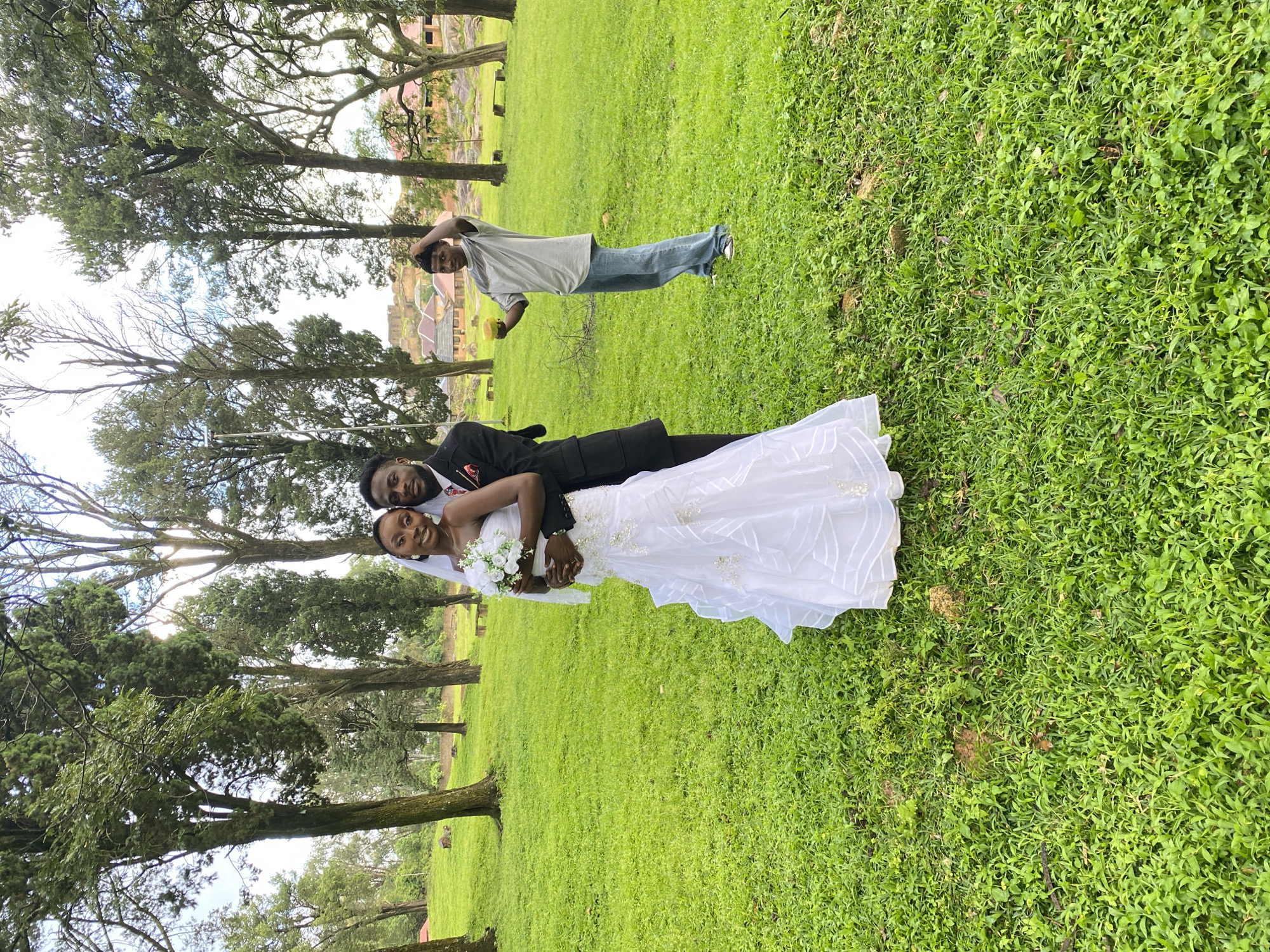 A full-shot, eye-level outdoor photograph captures a wedding ceremony in a grassy park. In the foreground, a bride and groom stand close together, the bride wearing a white, strapless wedding gown with a veil, and the groom in a black suit and tie. The bride holds a white bouquet. Behind them, a young man in a grey t-shirt and blue jeans stands to the right, holding a yellow object. The park setting features numerous tall trees with green foliage, a stone building in the background, and more trees and a bench visible further back. The ground is covered in lush green grass. The location is the Rayfield Resort in Jos, Nigeria.