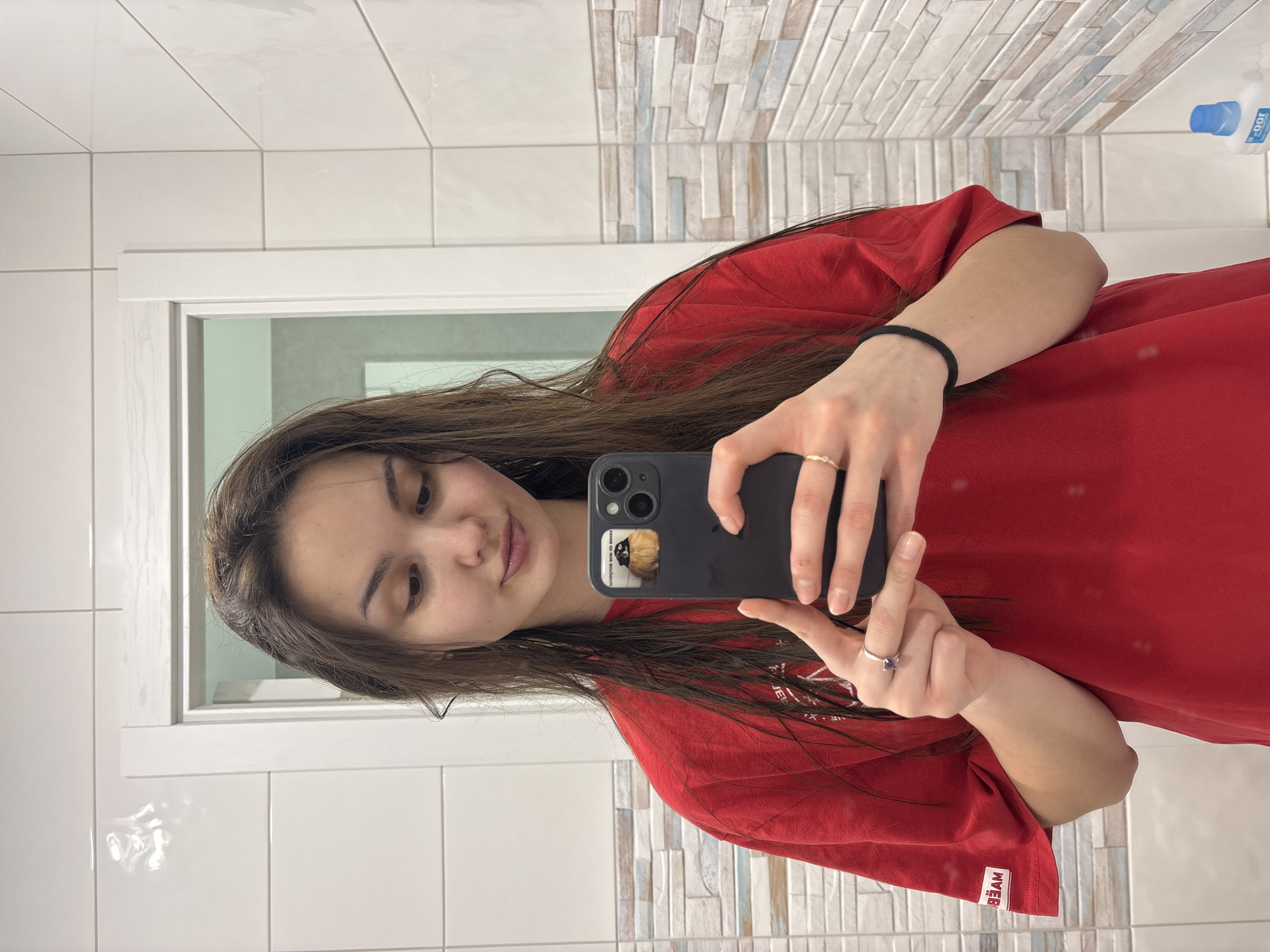 A young female is captured in a mirror selfie within what appears to be a bathroom in Chaikovsky, Russia. Her dark brown hair is wet and hangs loosely around her shoulders. She is wearing a red short-sleeved t-shirt. Her right hand holds a dark-colored smartphone, equipped with a sticker depicting a small, fluffy animal positioned above the camera array. A gold-toned ring with a clear stone is visible on her right ring finger, and a black hair tie encircles her left wrist. Her gaze is directed downwards towards the smartphone screen. The background features white rectangular wall tiles, with a horizontal accent band of varied rectangular tiles in shades of white, beige, light blue, and grey. A white door frame is visible in the upper left, revealing a light-colored room beyond. In the lower right corner, a white bottle with a blue cap rests on a surface, partially reflected.