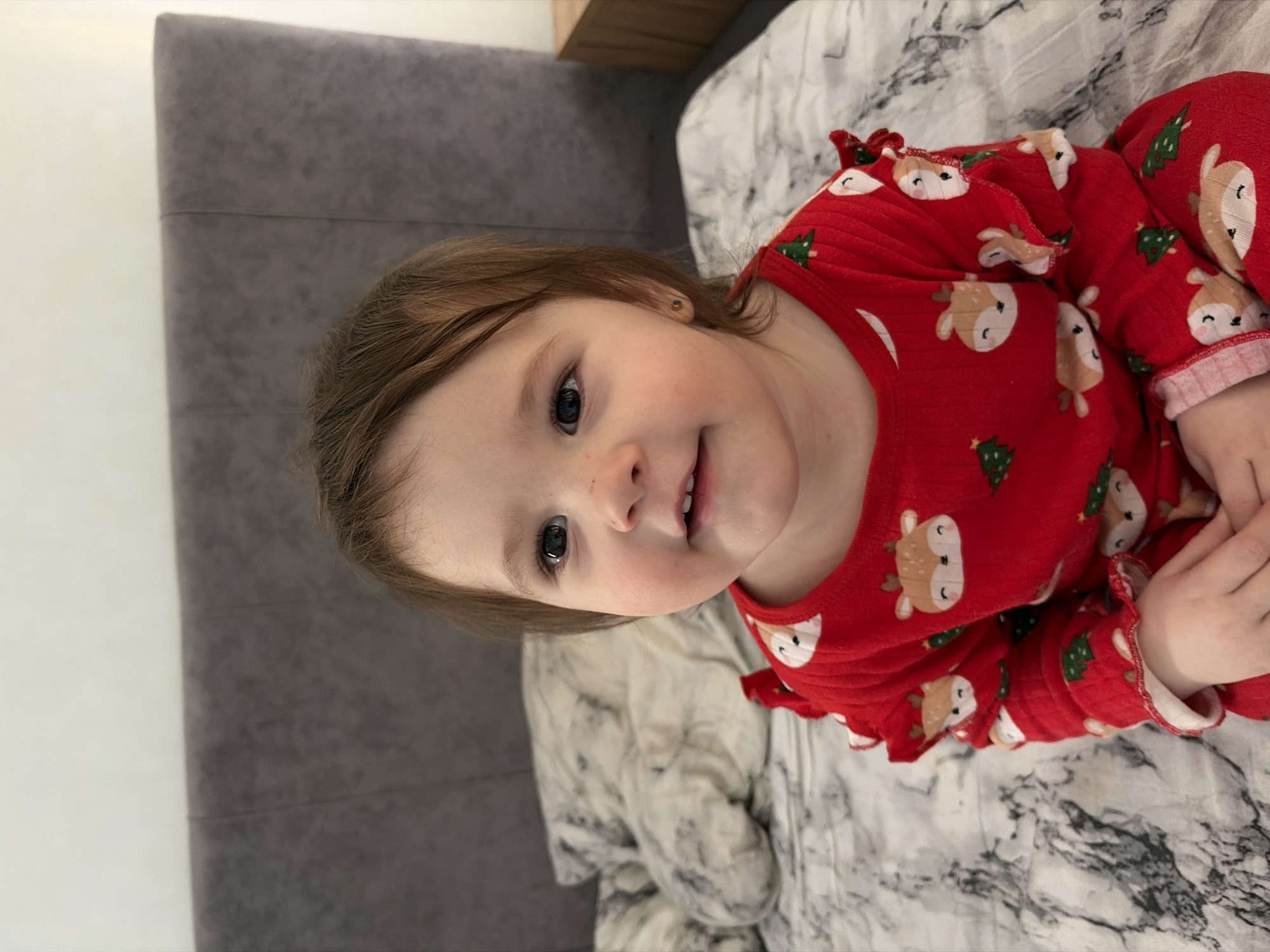A toddler wearing a red long-sleeved pajama top with reindeer and Christmas tree patterns lies on a bed covered with a gray and white patterned blanket. The bed is positioned against a gray headboard and a white wall. The toddler has light brown hair, fair skin, and blue eyes. The location is Chaikovsky, Russia.