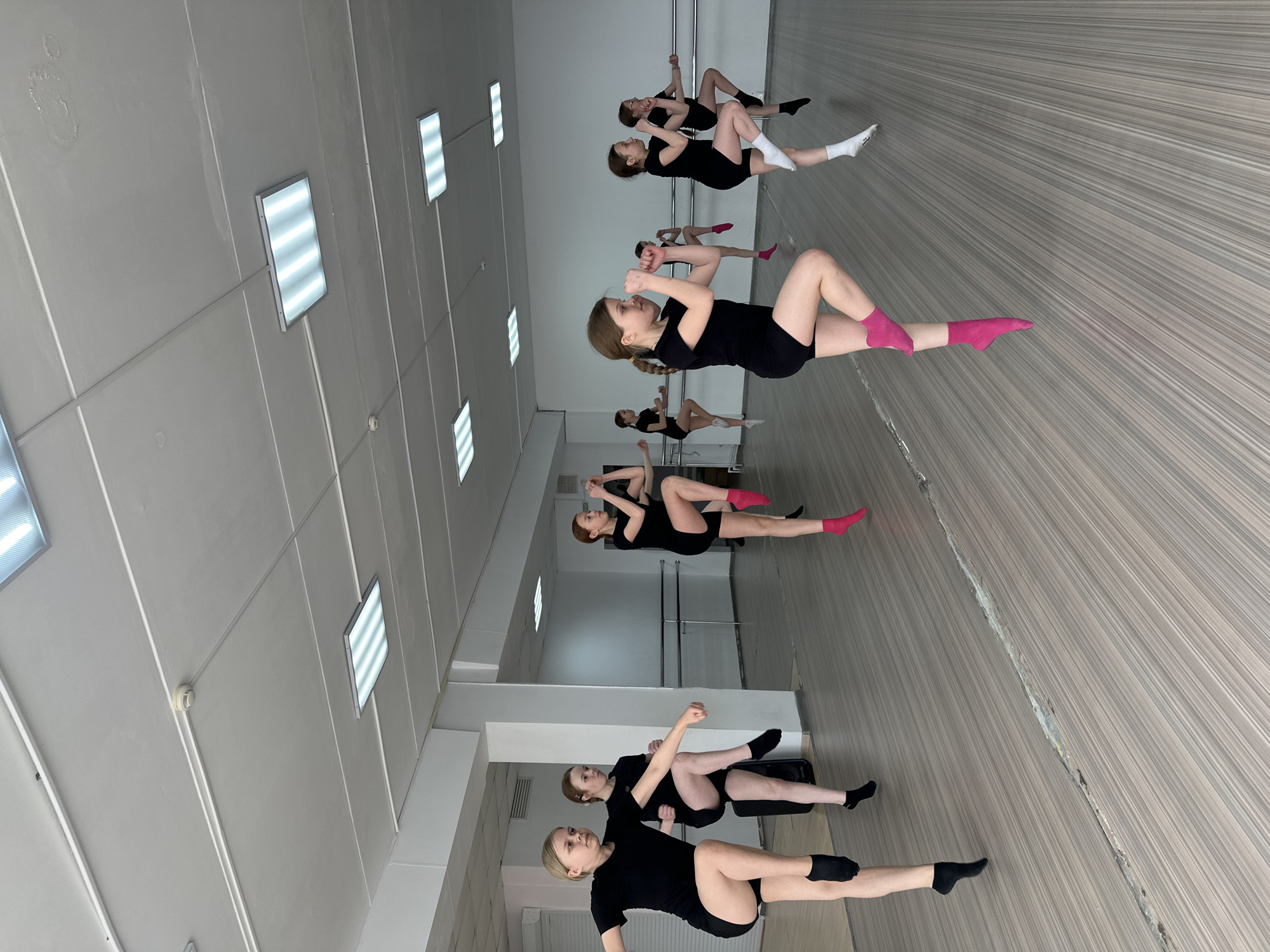 The image, taken in Votkinsk, Russia, depicts a group of approximately seven to eight young women or girls engaged in a floor exercise session within an indoor studio. The original image is rotated, showing the subjects as if on a vertical plane, but they are lying on their backs on the floor.
The participants are dressed uniformly in black athletic shorts and t-shirts. They are performing an exercise with bent knees and their hands positioned near their faces or chests, resembling crunches or a core workout. Some individuals wear brightly colored socks, specifically pink, while others have white or black socks.
The setting is a dance or fitness studio, characterized by light-colored, linear-grained laminate or wooden flooring. The floor shows visible seams and in places, a notable gap or crack. Mirrored walls reflect some of the individuals, and dance barres are mounted along the perimeter. The ceiling consists of white panels fitted with multiple rectangular fluorescent light fixtures, providing strong illumination. A smoke detector is visible on the ceiling, along with some irregular marks or stains on the panels, possibly indicative of water damage. No discernible text is visible in the image.