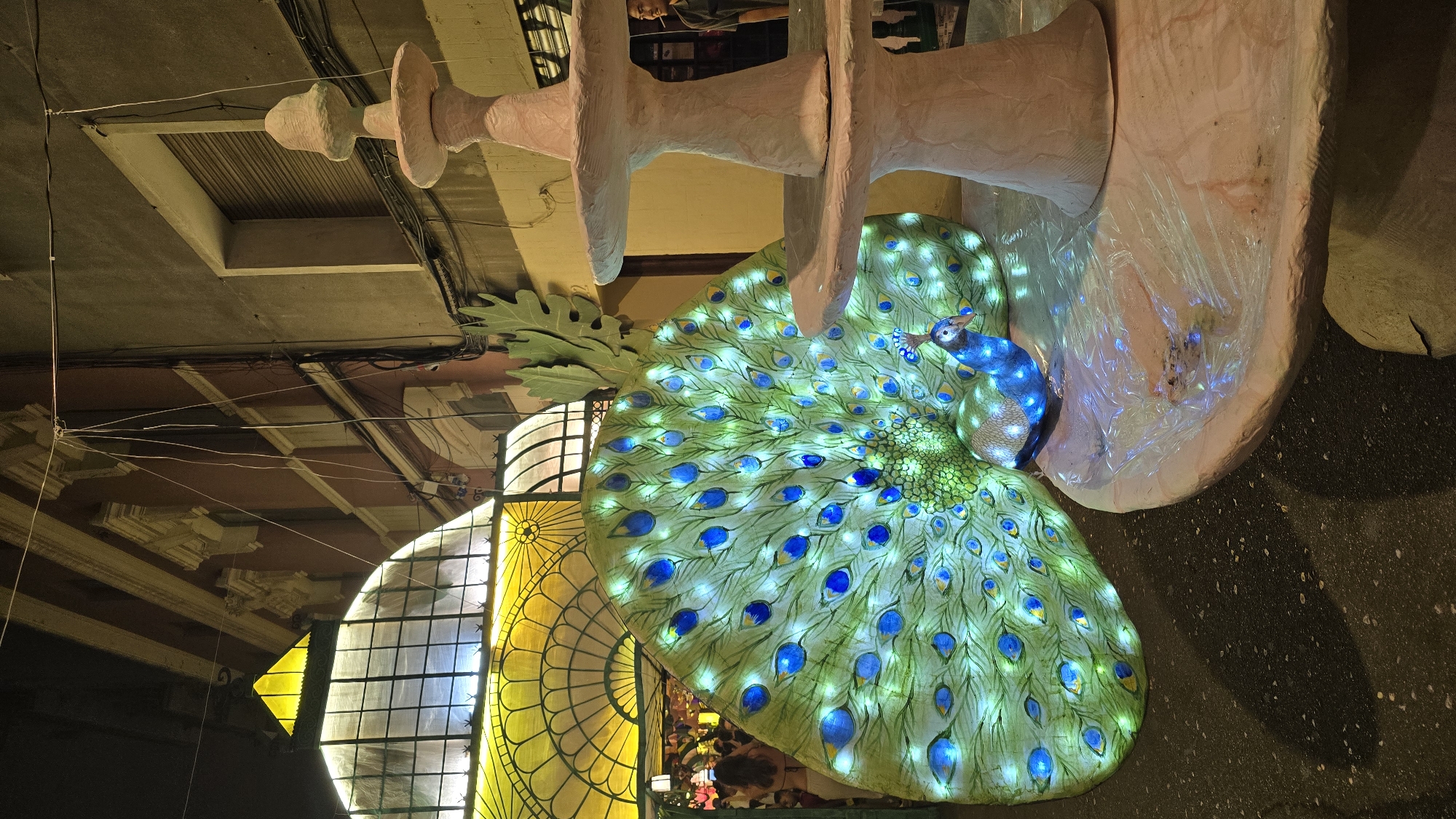 This is a nighttime outdoor shot of a decorative peacock lantern display, likely part of a festival or event in Barcelona, Spain. The peacock, illuminated by blue and green lights, is positioned in front of a large, fanned-out tail made of fabric or similar material, also lit with white and blue LEDs to mimic peacock feathers. The peacock is situated on a tiered, pinkish-beige fountain structure, covered in places with clear plastic sheeting. To the left of the peacock display is an ornate, arched entryway with yellow stained glass panels, behind which glimpses of people and more lights are visible. The background consists of buildings with balconies and electrical wires strung overhead. The ground is a dark, textured surface, possibly asphalt or pavement, with scattered reflections of light. A man in a dark shirt is visible to the far right, standing near the fountain. The scene appears to be set on a narrow street or plaza.