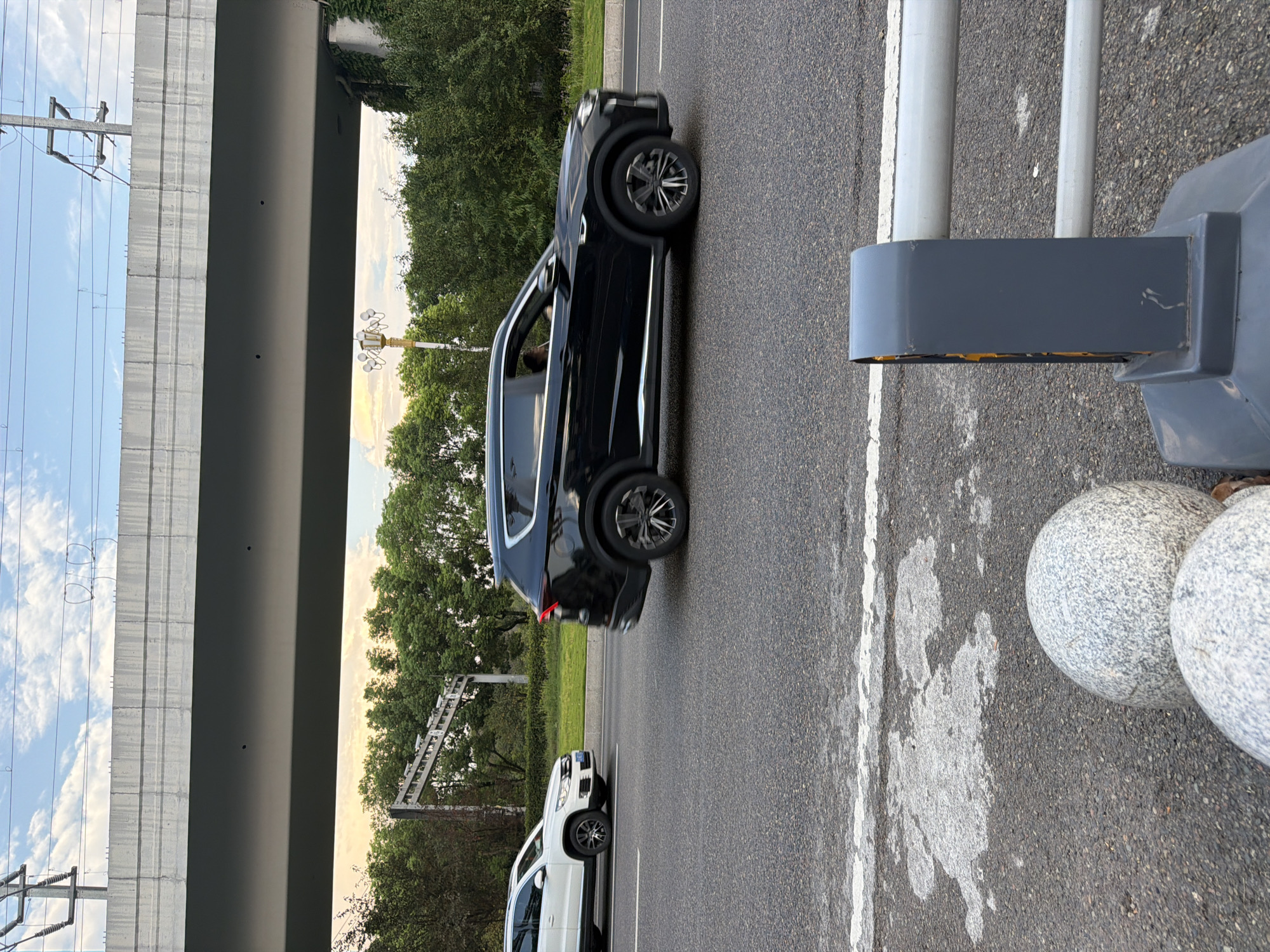 In Jiangjin, China, on a paved road under an elevated highway, a black SUV is driving on the right side of the road, moving from left to right. A white SUV is driving in the opposite direction on the left lane. The sky is partly cloudy with a gradient of colors from yellow to blue. Green trees line the side of the road behind the white SUV, and ivy grows on a concrete support structure of the elevated highway. In the foreground, a grey barrier with orange lights is visible, and two large, spherical stone bollards are positioned next to it. The road has white lane markings.