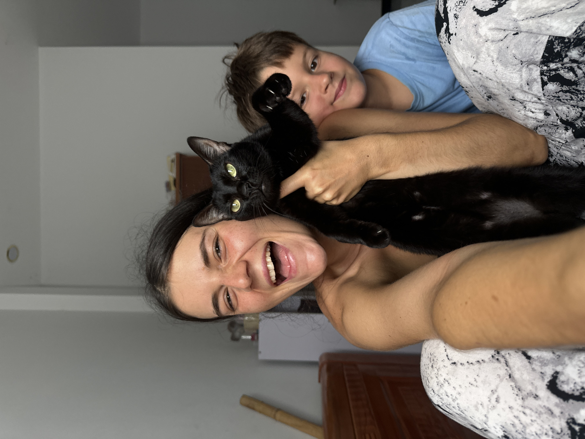 A young woman with dark hair is holding a black cat, with its head and paws visible. She is looking at the camera and laughing, with her mouth wide open. To her right, a young boy in a blue t-shirt is smiling. The setting appears to be indoors, with a plain wall and a piece of furniture visible in the background. The woman is wearing patterned pants. The scene is likely from Mũi Né, Vietnam, given the provided context.