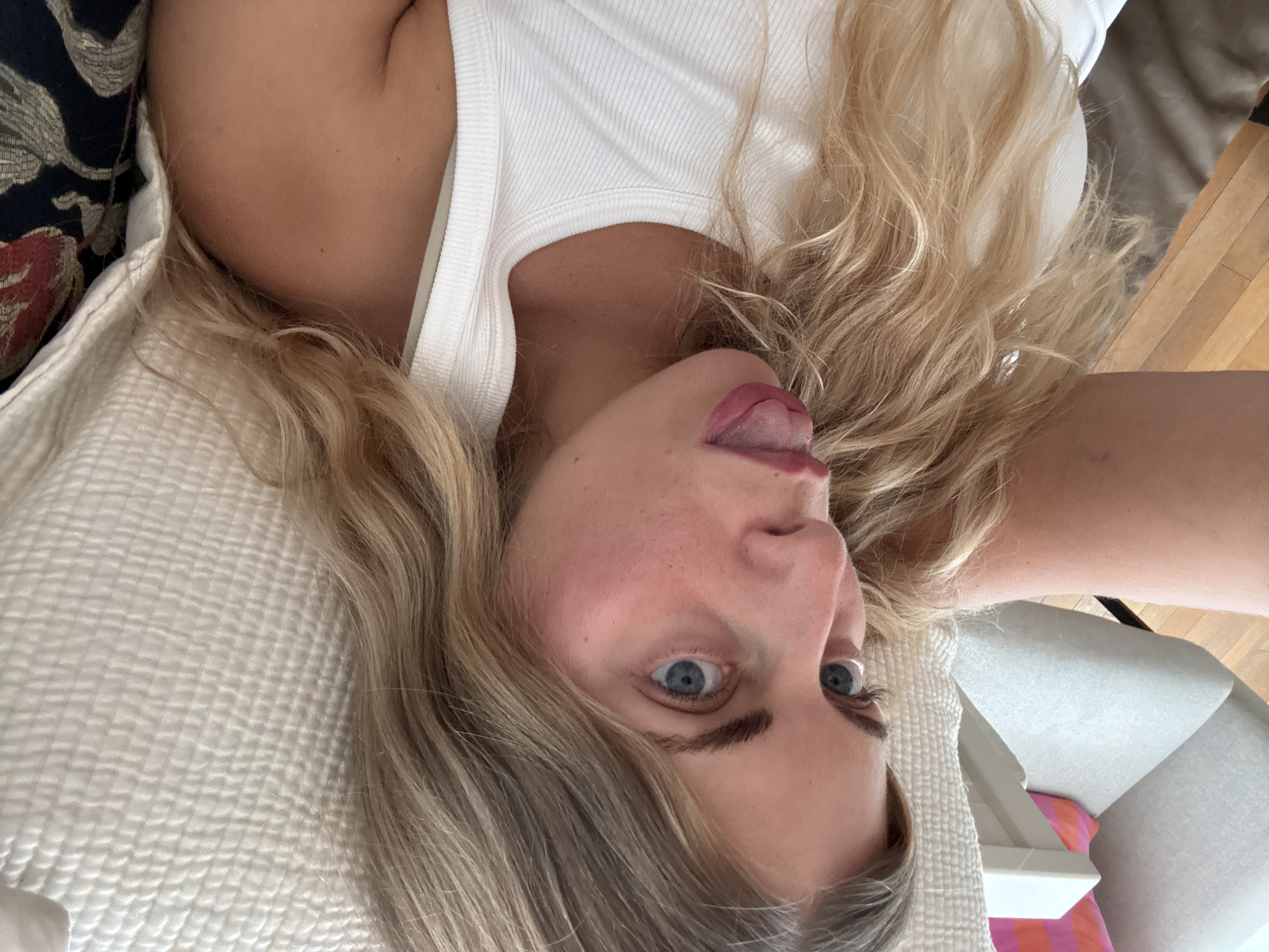 The scene is located in the city of Paris, France. The image features a close-up, eye-level perspective of a fair-skinned woman with long, wavy blonde hair and blue eyes. She is positioned diagonally within the frame, with her body angled to the left and her gaze directed off-center to the left. Her tongue is extended from her mouth in a playful or expressive gesture. She has visible freckles on her face and cheeks, and her eyebrows are dark and defined. She is wearing a white ribbed tank top, and her left shoulder and upper arm are partially visible in the foreground. The background consists of a domestic interior. To the immediate right, a light-colored, quilted textile, likely a pillow or blanket, is visible. Further to the right, a section of a dark-colored cushion with ornate red and gold patterns is present. To the left, a light grey or off-white armchair is partially visible, adorned with a pink and orange striped pillow. A wooden floor can be seen in the upper left background behind the armchair.