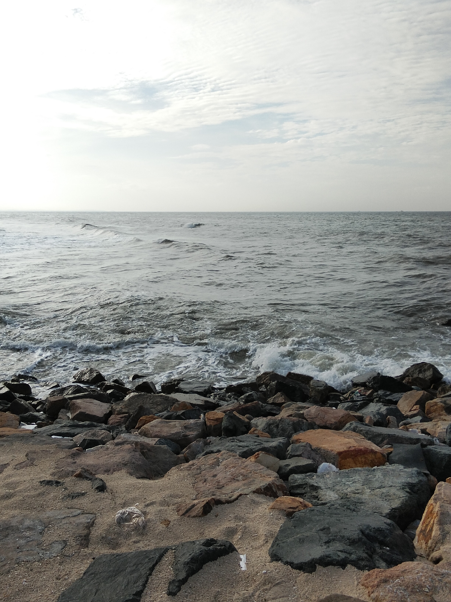 The image depicts a rocky shoreline with the ocean in the background. The sky is overcast with scattered clouds, and the sea is choppy with small waves breaking near the shore. Large, dark rocks are scattered along the edge of the water, and the foreground consists of sand and smaller rocks. There are no people or discernible man-made structures in the image, suggesting a natural, coastal environment. The scene is likely a beach or a rocky coast within the city of Phú Thủy, Vietnam.