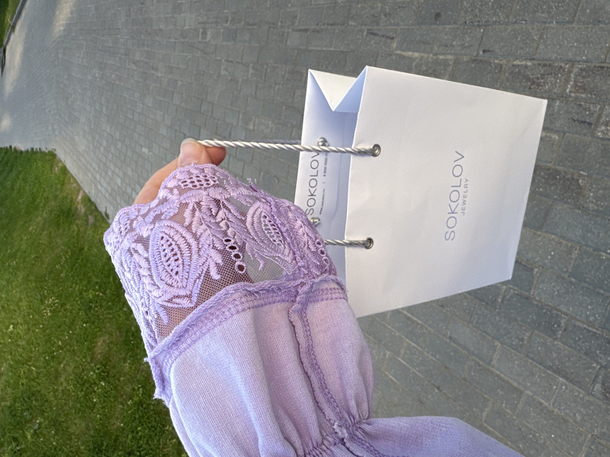 A close-up shot focuses on a person's left hand and arm as they hold a white paper shopping bag from "SOKOLOV JEWELRY." The hand is visible in the upper right quadrant of the image, grasping the bag's twisted white rope handle. The person is wearing a light purple long-sleeved garment with intricate lace detailing on the cuff, which is also visible in the foreground. The shopping bag features the "SOKOLOV JEWELRY" logo in grey text. The background consists of a paved walkway made of grey rectangular bricks, with a patch of green grass to the left, suggesting an outdoor urban or suburban setting. The scene is illuminated by natural daylight. The location is identified as Anudinovka, Russia.