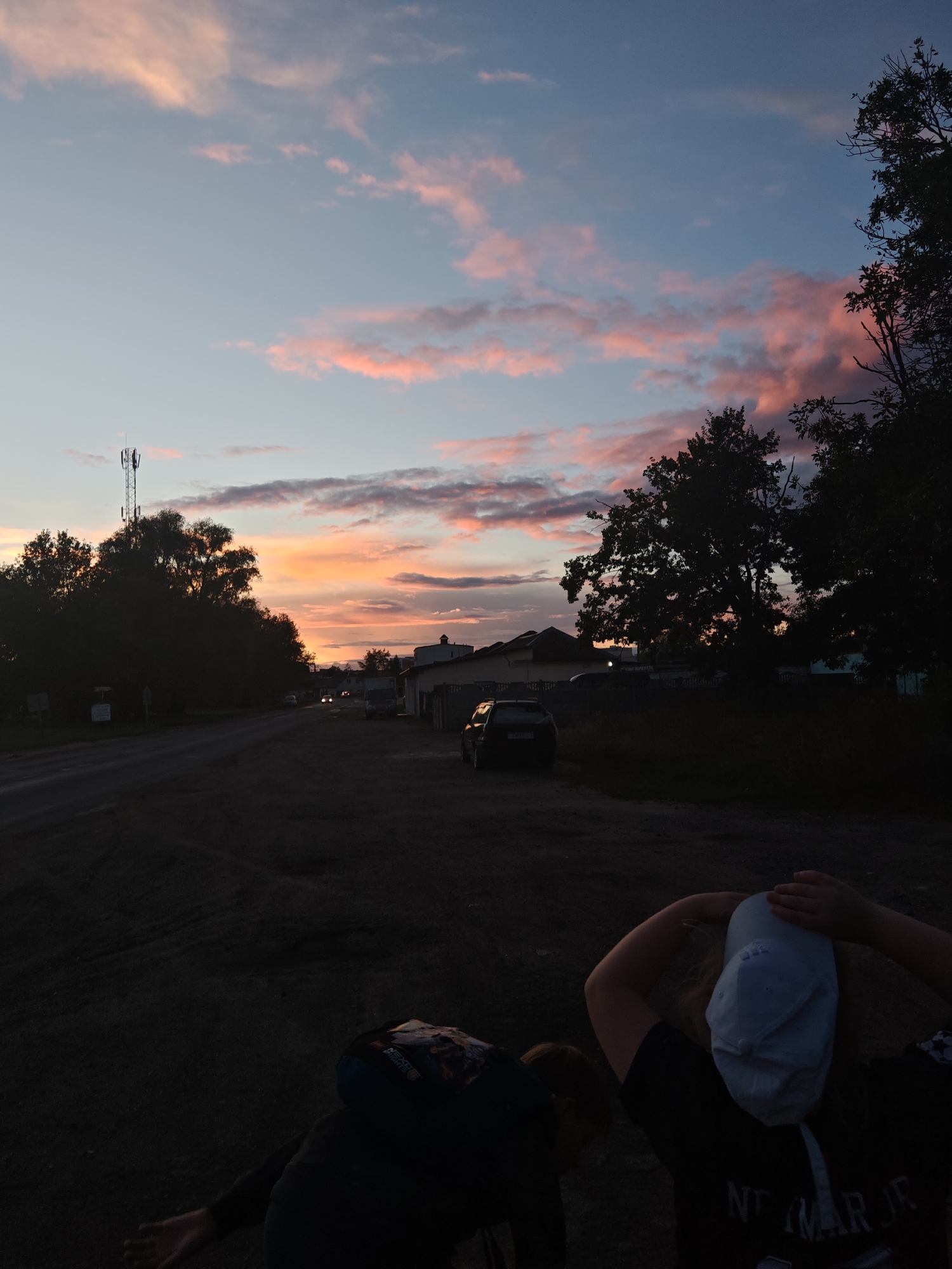 The image depicts a rural roadside scene at dusk. The sky is colored with hues of pink and orange from the setting sun, with scattered clouds. In the foreground, two individuals are partially visible. One person, wearing a dark hoodie and a baseball cap, has their back to the camera and is looking towards the sky. Another person, to the left, is also facing away from the camera. A dirt road stretches into the distance, lined with trees on either side. A telecommunications tower is visible in the background to the left, and several buildings and vehicles are scattered along the road. The overall atmosphere is tranquil and serene.