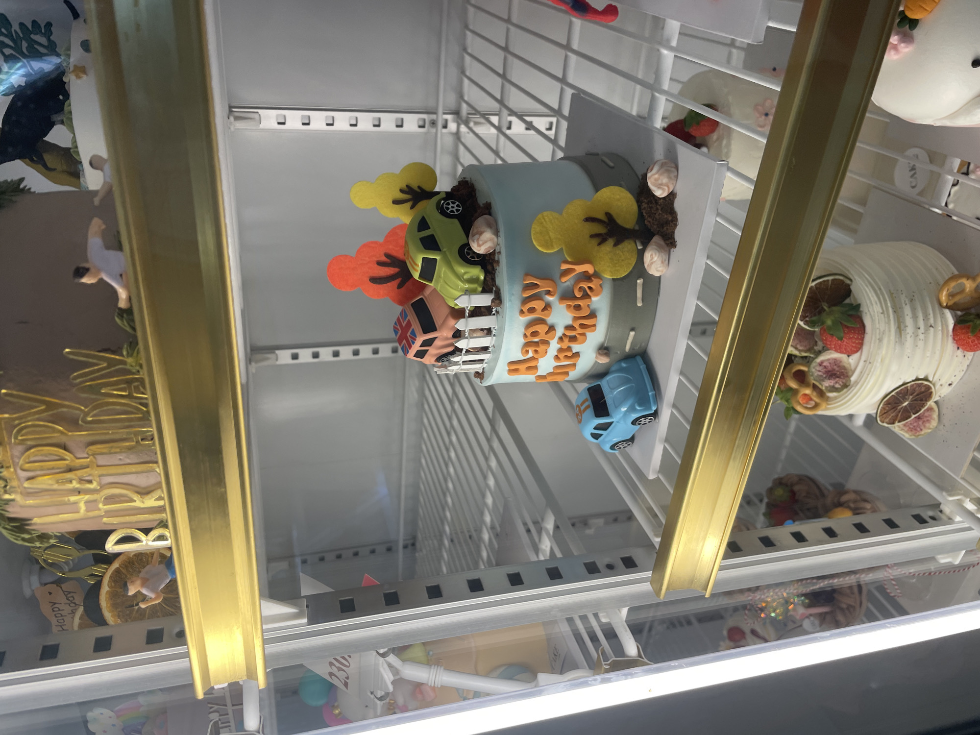 The image displays a bakery display refrigerator, likely located in Nha Trang Ward, Vietnam, filled with various cakes and desserts. Prominently featured is a light blue, cylindrical cake decorated with orange lettering that reads "Happy birthday." This cake is adorned with miniature toy cars, a white picket fence, and felt trees, suggesting a child-oriented theme. Further down, another cake is visible, this one white and decorated with fruits such as strawberries and dried citrus. The refrigerator shelves are made of white wire, and the interior lighting is a bright, cool white. The overall impression is one of a well-stocked, clean establishment showcasing baked goods.