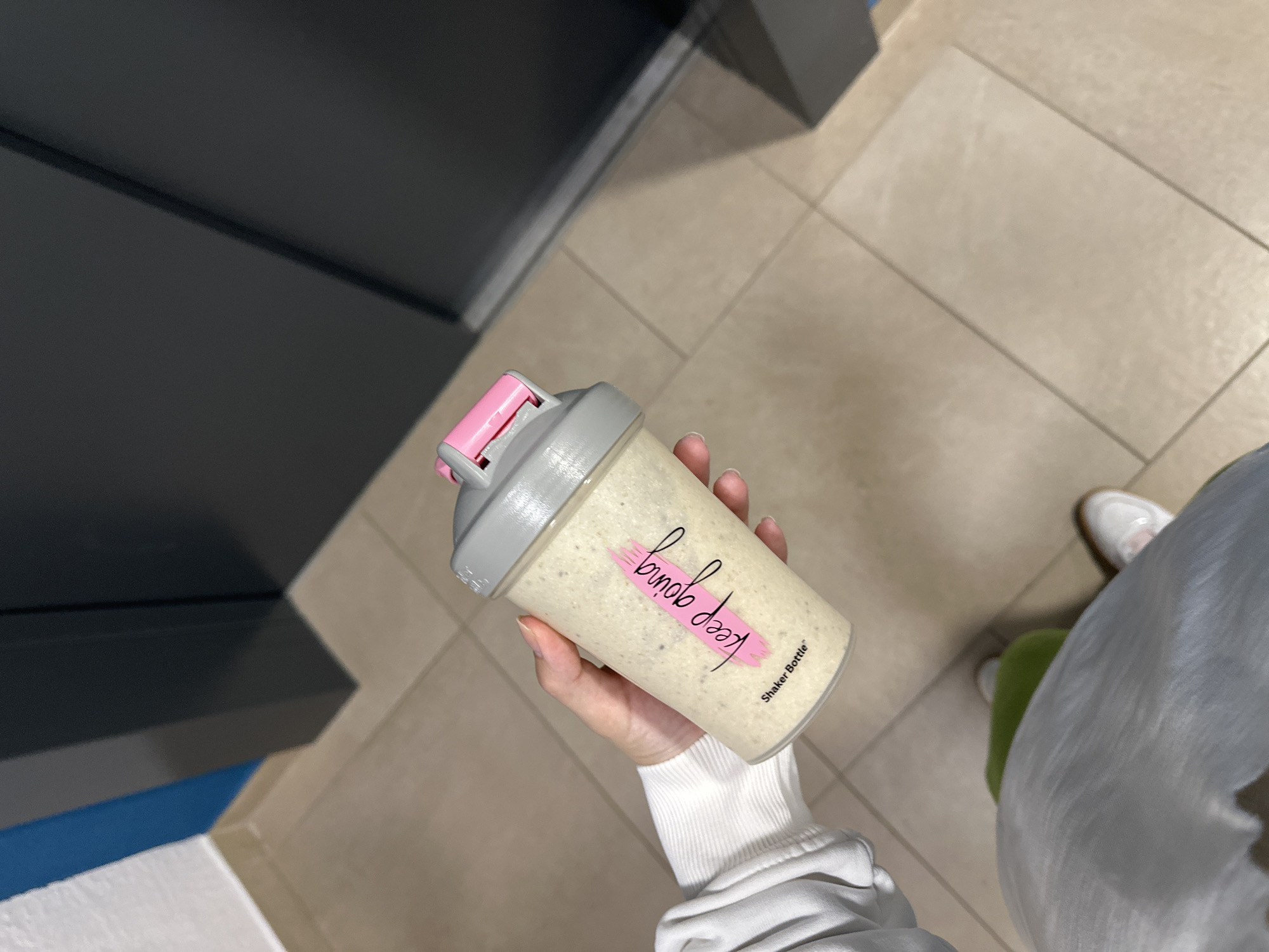 A partial view of an individual in Perm, Russia, holding a transparent plastic shaker bottle filled with a beige, speckled liquid. The bottle features the text "keep going" in a pink brushstroke script and "Shaker Bottle™" in smaller black text, and is topped with a grey screw-on cap that has a pink flip-top spout. The individual's right hand, with manicured nails, holds the bottle centrally. Visible clothing includes the ribbed cuff of a white long-sleeved top, light grey athletic pants, and a white sneaker with a light brown sole on the right foot, positioned near a partially visible bright green object on the floor. The setting is an indoor space with light beige rectangular floor tiles and dark grout lines. The background includes a corner where a white wall base transitions to an electric blue upper section, separated by a thin grey vertical trim, leading to a dark grey vertical panel or door structure on the right side of the frame.