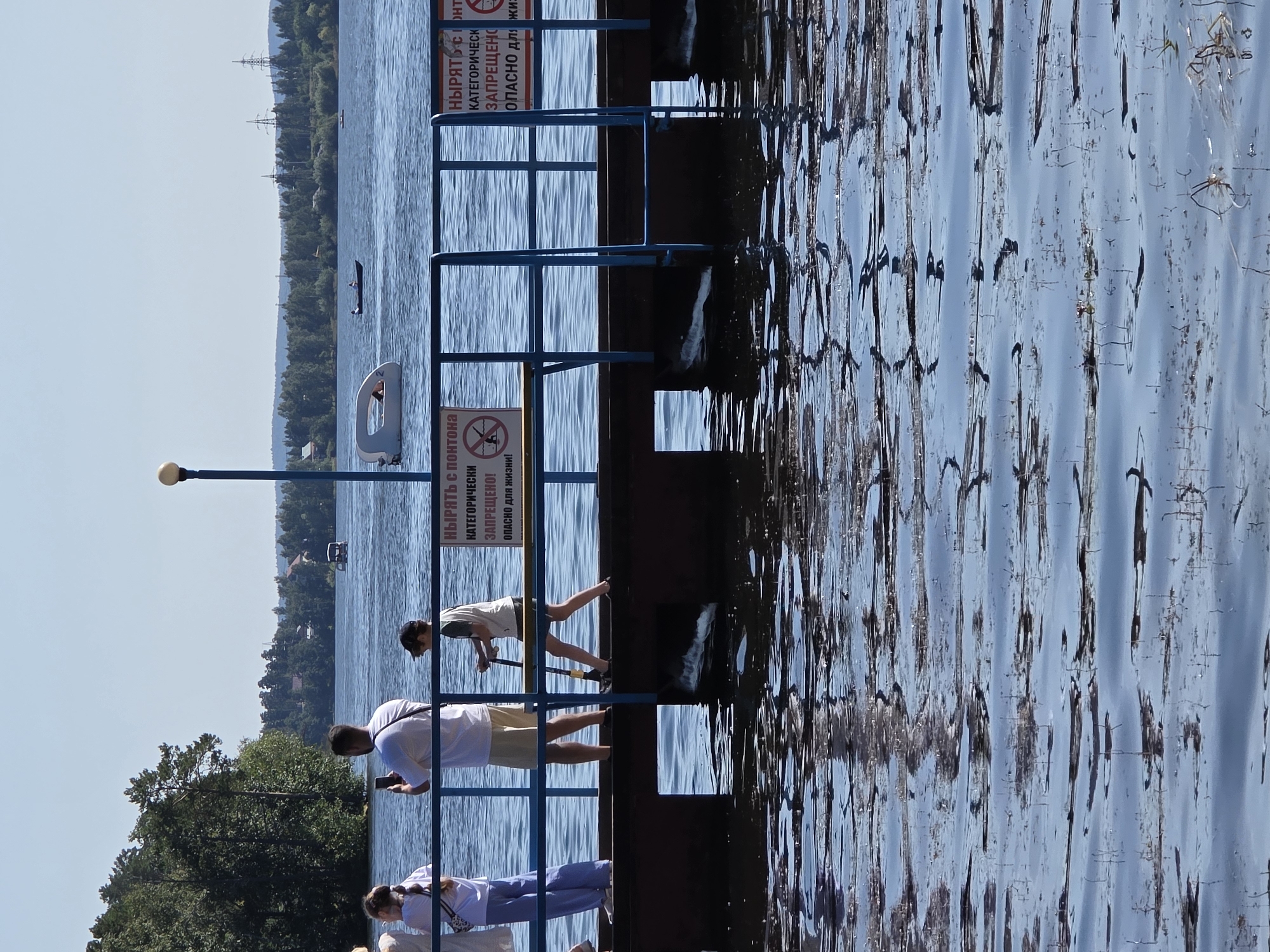This image depicts a sunny day on Lake Chebarkul in Chebarkul, Russia. In the foreground, the water is calm with reflections of the pier and surrounding vegetation. A wooden pier extends into the lake, with a blue railing along its edge. Several people are present on the pier. On the left, two young women and a man are standing. The man is holding a smartphone, appearing to take a picture. Further along the pier, a young boy wearing a cap and t-shirt is holding a long pole, possibly for fishing or some other activity. On the pier, there are two signs with Russian text. One reads "Нырять с понтона категорически запрещено! Опасно для жизни!" which translates to "Diving from the pontoon is strictly forbidden! Dangerous to life!". Another partially visible sign carries a similar warning. In the background, across the lake, a treeline and some buildings are visible. Several boats are on the water, including a small white watercraft with a person inside and a darker boat with two occupants. A few utility poles are also visible in the distance. The overall scene is one of a recreational area by a lake on a clear day.