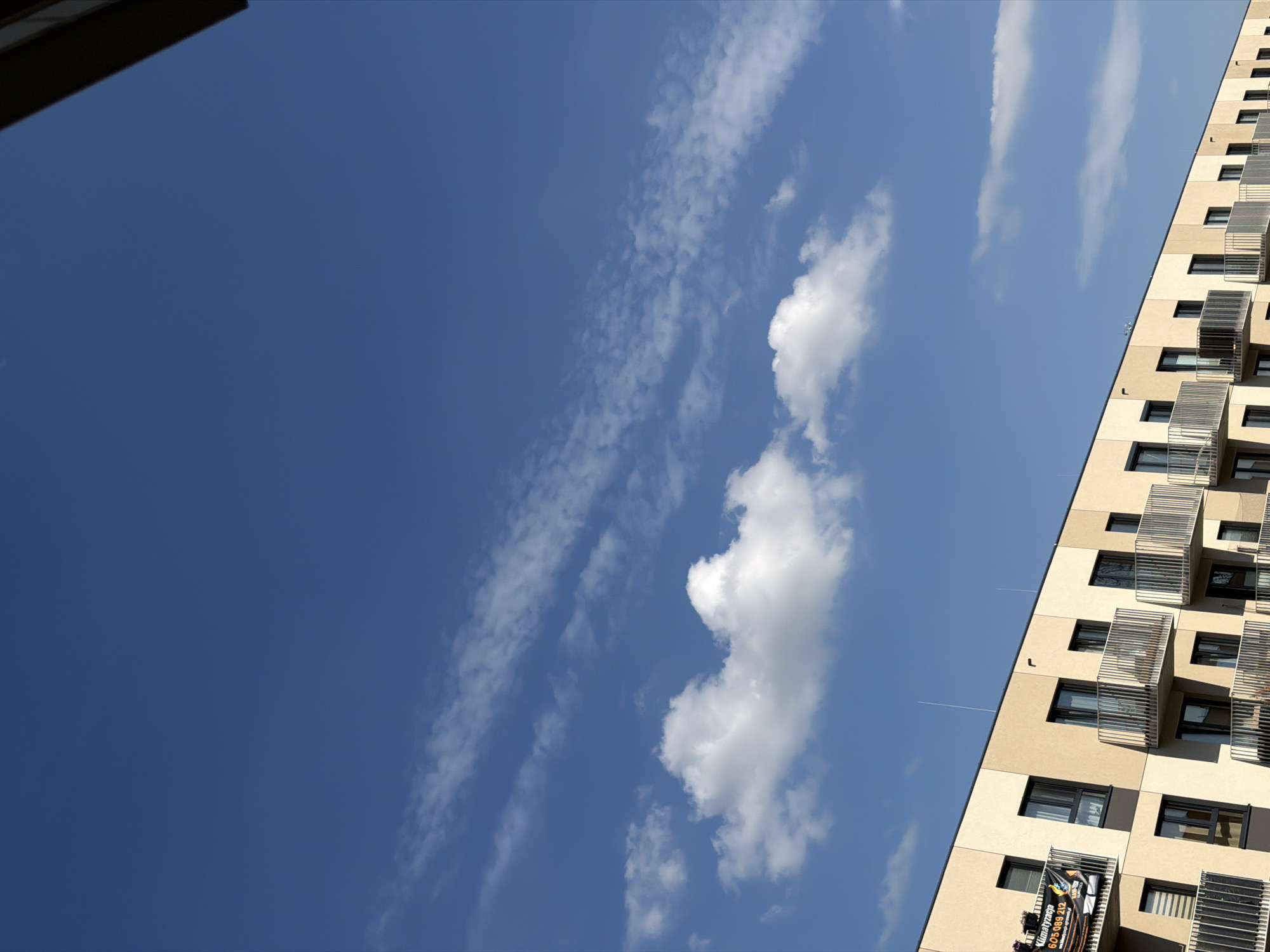 The image shows a modern apartment building with beige and dark gray facades, featuring numerous balconies with metal railings. The building is located in the city of Łódź, Poland. The sky is clear blue with scattered white clouds, some of which are wispy and stretched across the frame. A banner with Polish text and advertisements is visible on one of the balconies on the left side of the building. The scene is captured from a low angle, emphasizing the building against the expansive sky.