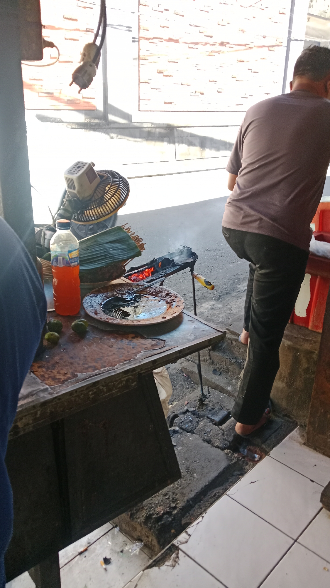 In the city of Kuta, Indonesia, an outdoor food stall displays a variety of items on a weathered wooden table. To the left, a person wearing a blue shirt is partially visible, facing away from the camera. In the center of the table, a clear plastic bottle filled with orange liquid stands next to a woven basket containing green banana leaves and what appear to be raw food items. In front of these items, a dark, circular plate holds a dark liquid. Further to the right, a portable grill with glowing red embers is positioned over a concrete surface. A person wearing a purple shirt and black pants stands to the right of the grill, facing away from the camera, with one foot visible on the ground. Behind the stall, a brick wall is visible, bathed in sunlight. The ground is a mix of concrete and dirt, with some scattered debris. A fan is visible on the left, behind the table.