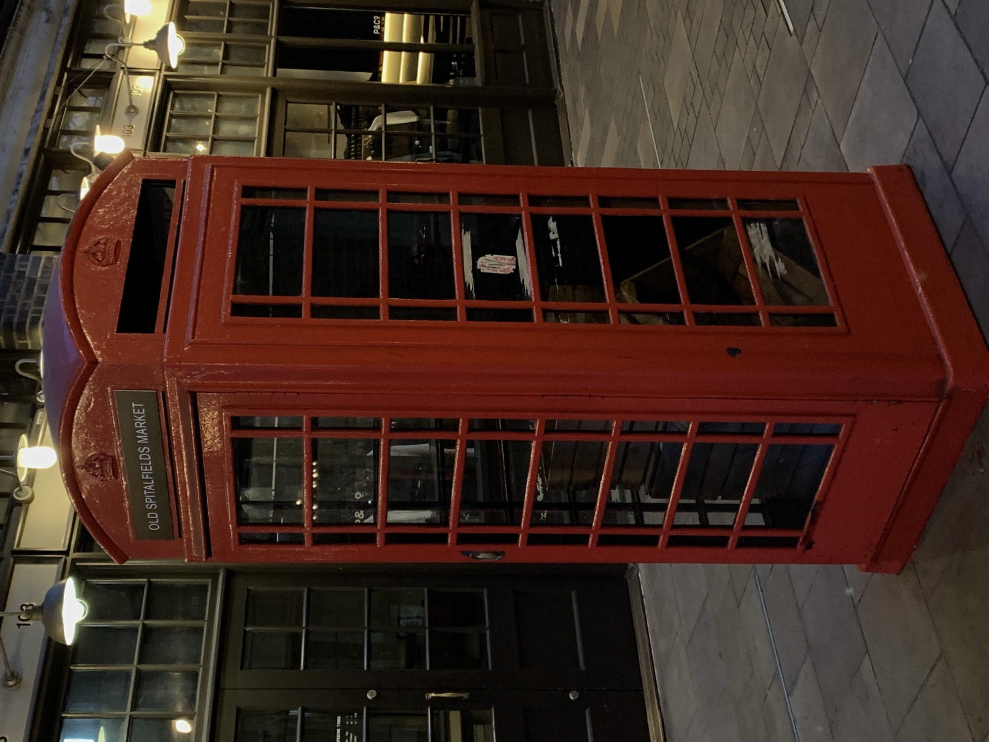 A classic red British telephone booth, specifically a K6 Kiosk model, stands prominently on a paved surface in an urban setting. The booth is slightly worn, with some visible imperfections on its red painted surface. Near the top of the booth, a Royal Cypher (a crown symbol) is embossed, and a black sign with white text clearly reads "OLD SPITALFIELDS MARKET". Inside the booth, a sticker or piece of paper is visible on one of the glass panes.
In the background, a multi-story building with numerous large, gridded windows stretches across the frame. The scene is illuminated by artificial lights attached to the building's facade, indicating it is evening or night. Through one of the upper windows, the text "P&C O" can be faintly discerned. Below these windows, signs on the building display the numbers "103" and "105", likely denoting addresses or unit numbers. The paved ground beneath the booth is visible, suggesting a street or market area. No individuals are present in the image. The iconic telephone booth and the "OLD SPITALFIELDS MARKET" text strongly suggest the location is London, United Kingdom.