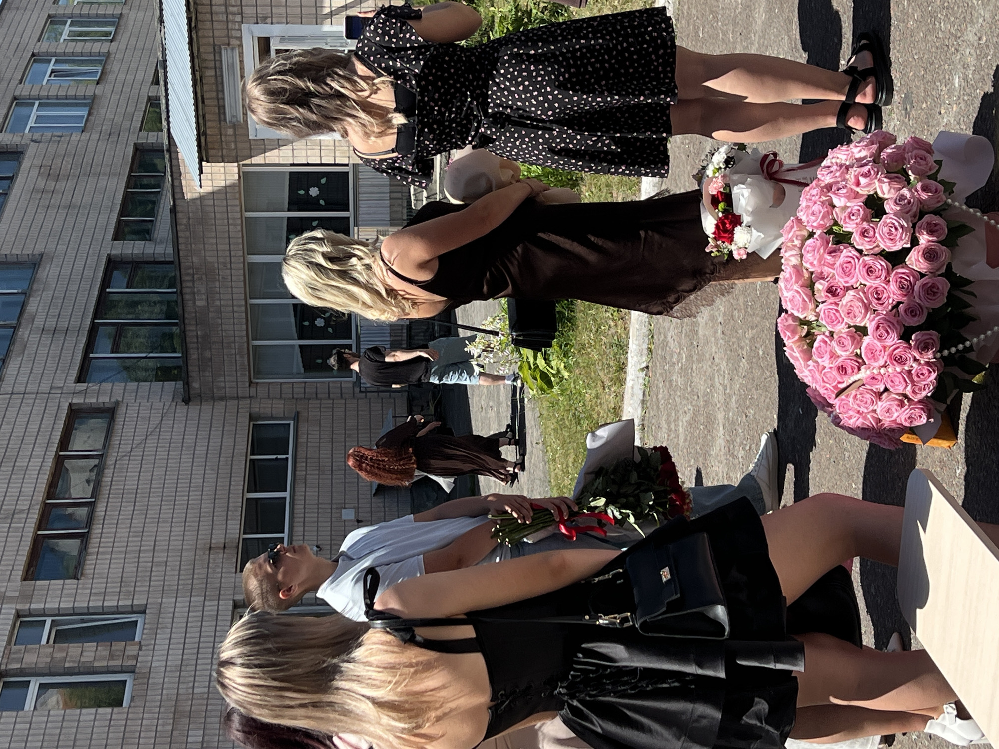 In Ternopil, Ukraine, several people are gathered outdoors near a brick building with multiple windows. The scene appears to be a sunny day, possibly in the afternoon, indicated by the strong shadows cast on the pavement.
The primary subjects are several young women, some of whom are holding large bouquets of flowers. One woman in a black dress with a white polka dot pattern is prominent, as is another in a dark-colored dress. A third woman with blonde hair wears a black outfit. Another individual, appearing to be a woman with red hair, is seen in a brown dress. A person in a light blue t-shirt and jeans is also visible.
The activity seems to be a celebration or a farewell, given the presence of the bouquets and the celebratory attire of some individuals. One large bouquet is composed of many pink roses, while another features red roses. A smaller bouquet with red and white flowers is also present. The individuals are standing on a paved surface, possibly a sidewalk or street, with a grassy area to the side of the building. The building itself is a multi-story structure made of light-colored bricks, featuring numerous rectangular windows.
One person is holding a mobile phone, possibly taking pictures or videos. The overall mood seems somewhat formal yet relaxed, with a hint of solemnity due to the flowers. No visible text is discernable in the image. The setting is urban, characterized by the building and paved ground.
