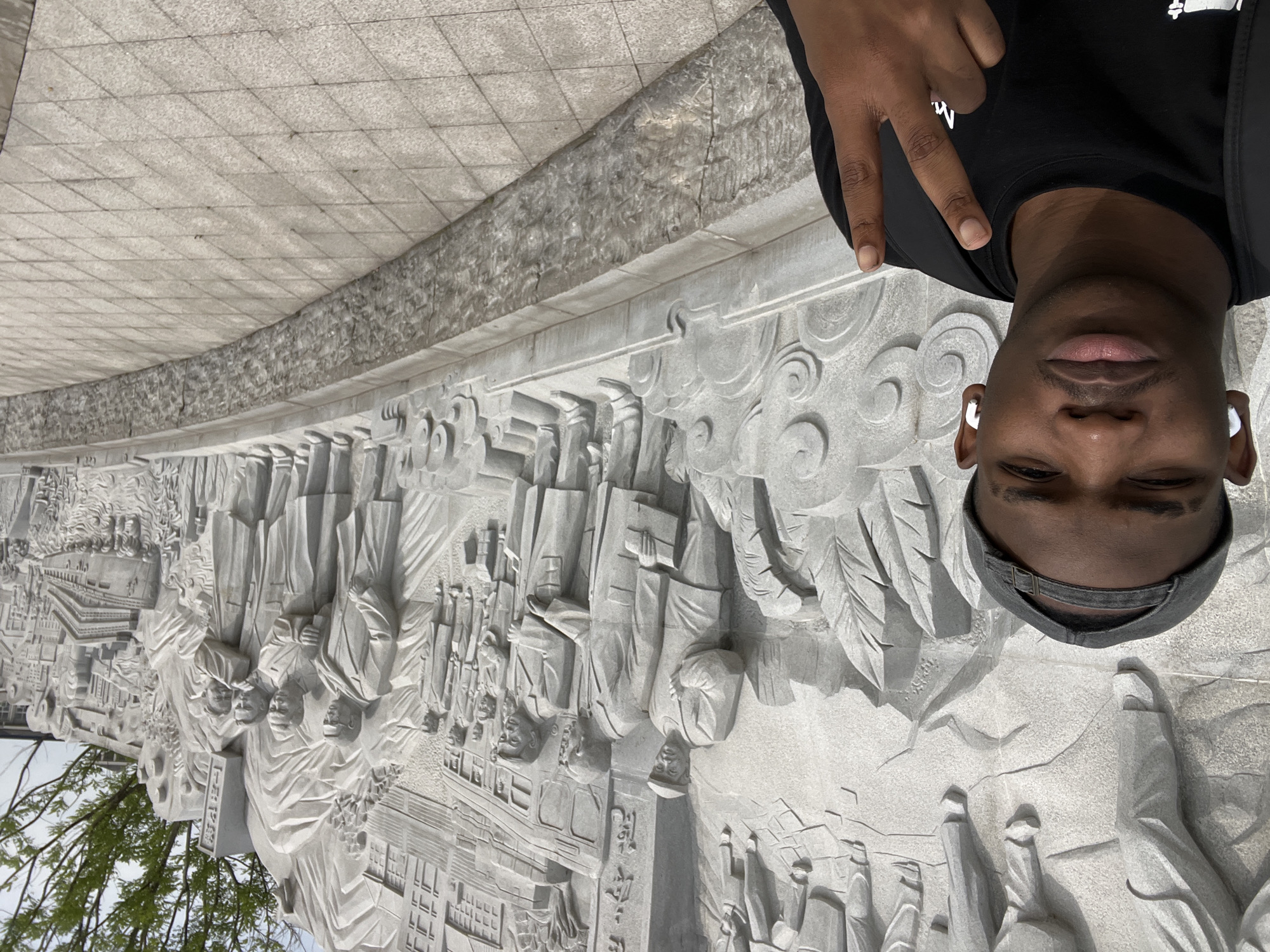 A man takes a selfie in front of a stone relief carving in Chongqing High-tech Zone, China. The carving depicts a historical scene with multiple figures in what appears to be traditional clothing or uniforms. The man is wearing wireless earbuds, a black T-shirt, and a baseball cap. He is holding up two fingers in a peace sign. The background shows a paved area and some greenery.