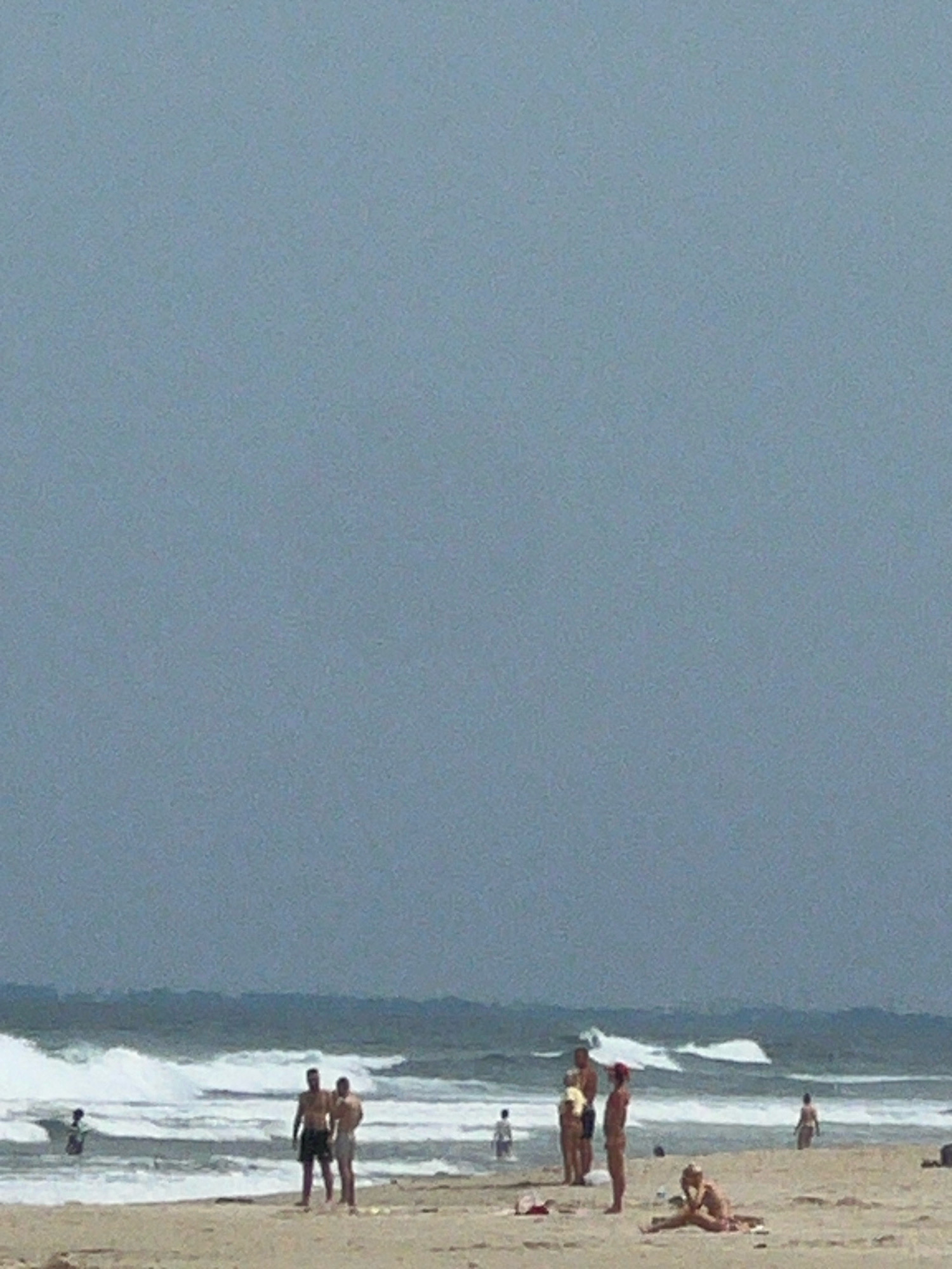 A cloudy day at a beach with gentle waves. Several people are visible, some standing in shallow water, others on the sand. Two men in swim trunks stand together on the shore, while a person in the distance appears to be swimming. Further down the beach, a woman sits on the sand, and another figure stands near her. The sky is overcast, and the water has a greyish hue. It is not possible to determine the specific location in Da Nang, Vietnam from the image.