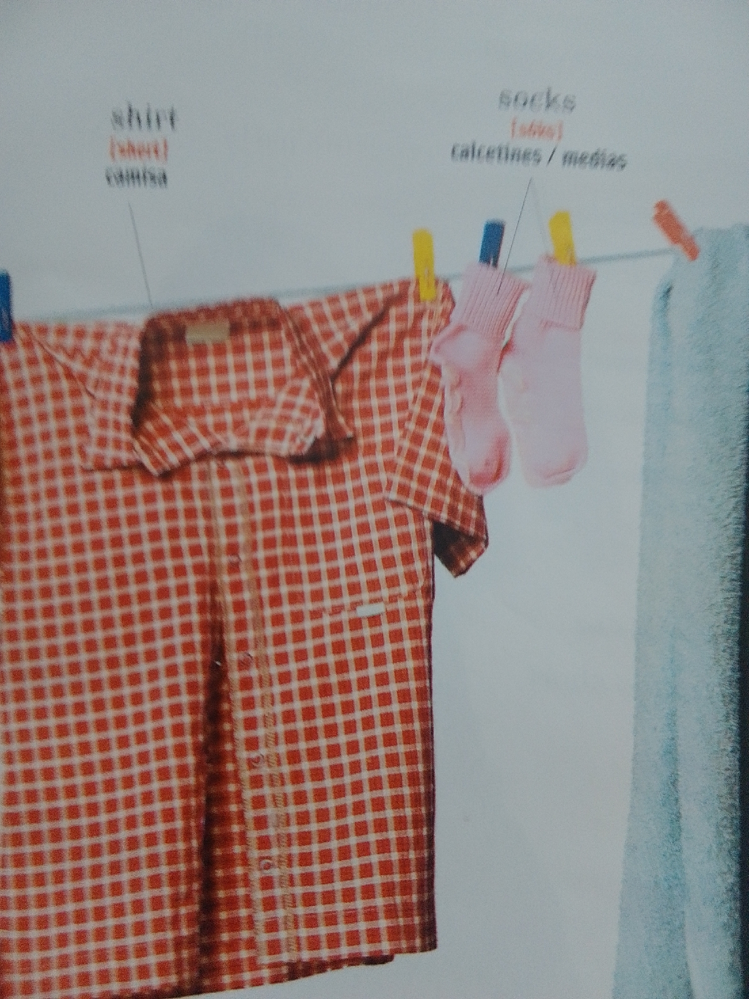 A red and white checkered shirt hangs on a clothesline, held by clothespins. Next to the shirt, two light pink socks are also hanging on the line with clothespins. A light blue towel or piece of fabric is visible to the right, also on the clothesline. The background is a plain, light-colored surface, likely a wall.
Text visible in the image includes: "shirt" with a line pointing to the shirt, and "(camisa)" and "shirt" in smaller text below it. To the right, "socks" is written with a line pointing to the socks, and "(socks)" and "calcetines / medias" in smaller text below it.
The scene depicts laundry drying outdoors. The lighting suggests daytime. The overall mood is domestic and ordinary.