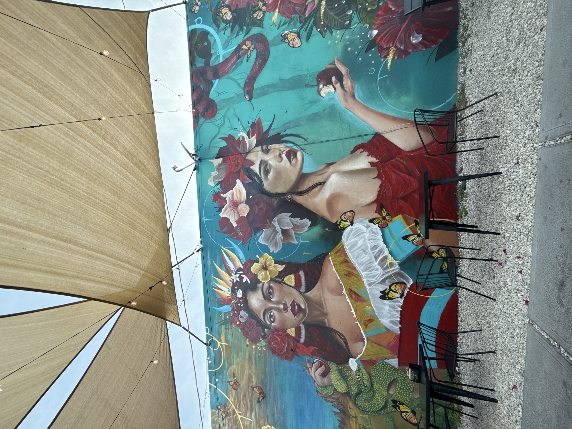 The image displays an outdoor patio scene dominated by a vibrant, large-scale mural on a wall, captured from a low angle. The mural features two ornately adorned women amidst a lush, symbolic landscape.
The upper woman in the mural wears a red, off-the-shoulder garment and an elaborate floral headpiece with red and white flowers. She has dark hair, dark eyes, and red lips, gazing intently upwards and to the right. Her right hand, raised, holds a bitten red apple. Above her, a red and black-banded snake coils against a teal background, which is dotted with large red hibiscus flowers, green foliage, and numerous yellow butterflies. Faint circular outlines also appear on the teal.
The lower woman in the mural is depicted wearing a white, puffy-sleeved top and a colorful, patterned dress. Her dark hair is crowned with a striking arrangement of yellow, red, and white flowers and feathers. She looks directly towards the viewer with an intense expression. Below her, a green snake with dark spots coils, and she holds another red fruit, possibly an apple, in her hand. More butterflies flutter around both figures. The mural's style is richly detailed and colorful, evoking a mythological or allegorical narrative, potentially referencing themes like nature, temptation, or femininity.
To the left of the mural, several light tan shade sails are stretched overhead, forming geometric patterns against a bright, clear sky. String lights with small, exposed bulbs are strung between the sails and along the edge of the mural wall. On the right side of the image, the ground is covered in light-colored gravel, with scattered small purple petals. Several black metal chairs with slatted backs and seats are arranged on the gravel, some appearing to be stacked. A concrete curb or divider borders the right edge of the frame.
The setting suggests a charming and artistic outdoor dining or lounge area, likely experienced during the daytime under pleasant weather. The location is Hollywood, United States of America. No visible text is present in the image.