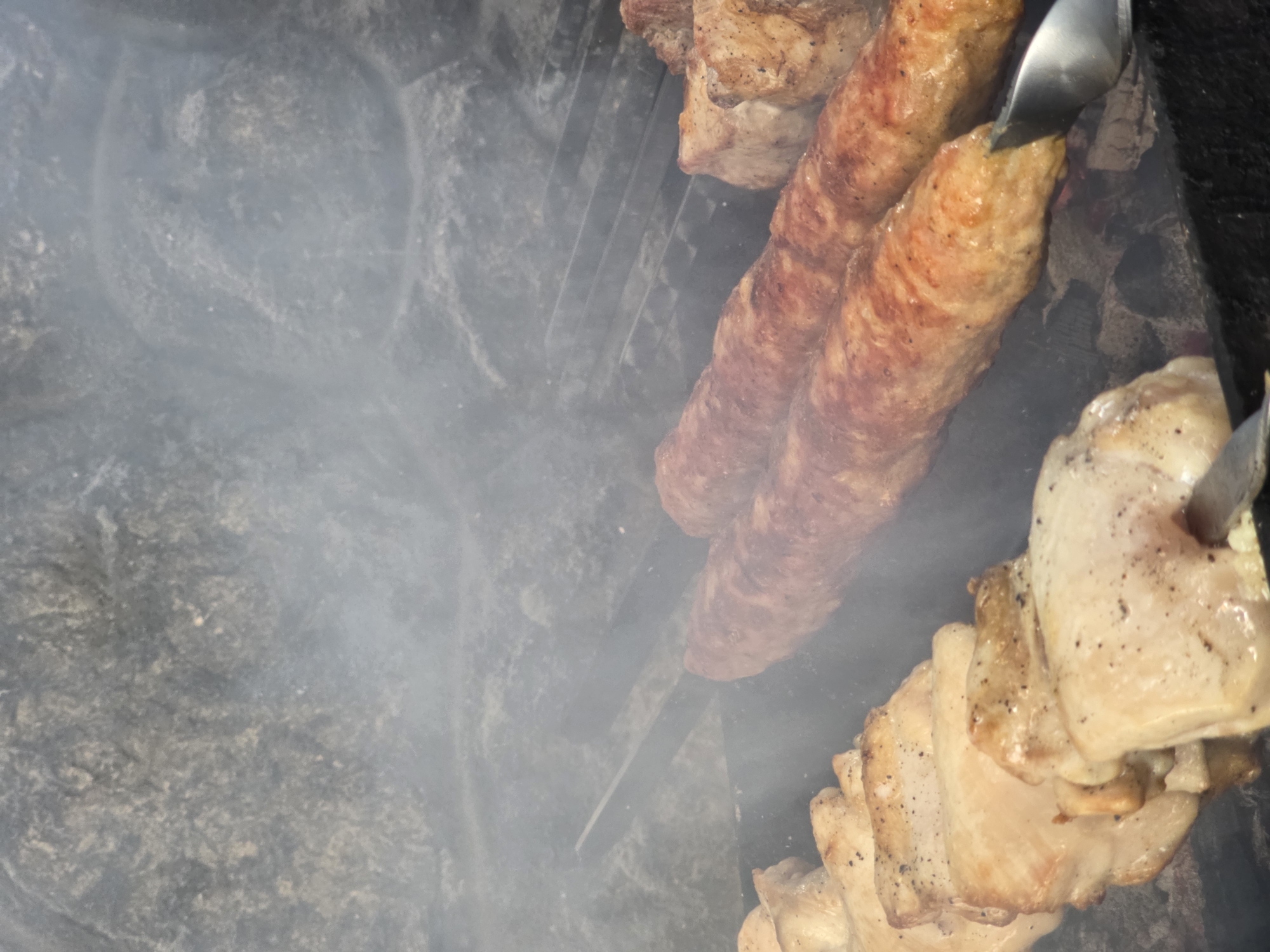 The media file shows a close-up, overhead view of food being grilled outdoors. The subjects are pieces of meat cooking on a grill. There are long, cylindrical sausages and pieces of what appear to be chicken or pork belly. Tongs are visible, holding and manipulating one of the sausages. Smoke is rising from the grill, obscuring parts of the scene. The background suggests a rustic setting, with rough, dark stone or earth visible. The lighting indicates it is daytime. The overall impression is of a barbecue or outdoor cooking event. There is no visible text. The location context is Pidhorodne, Ukraine, suggesting a possible outdoor gathering or a meal prepared in a rural or semi-rural setting in that region.