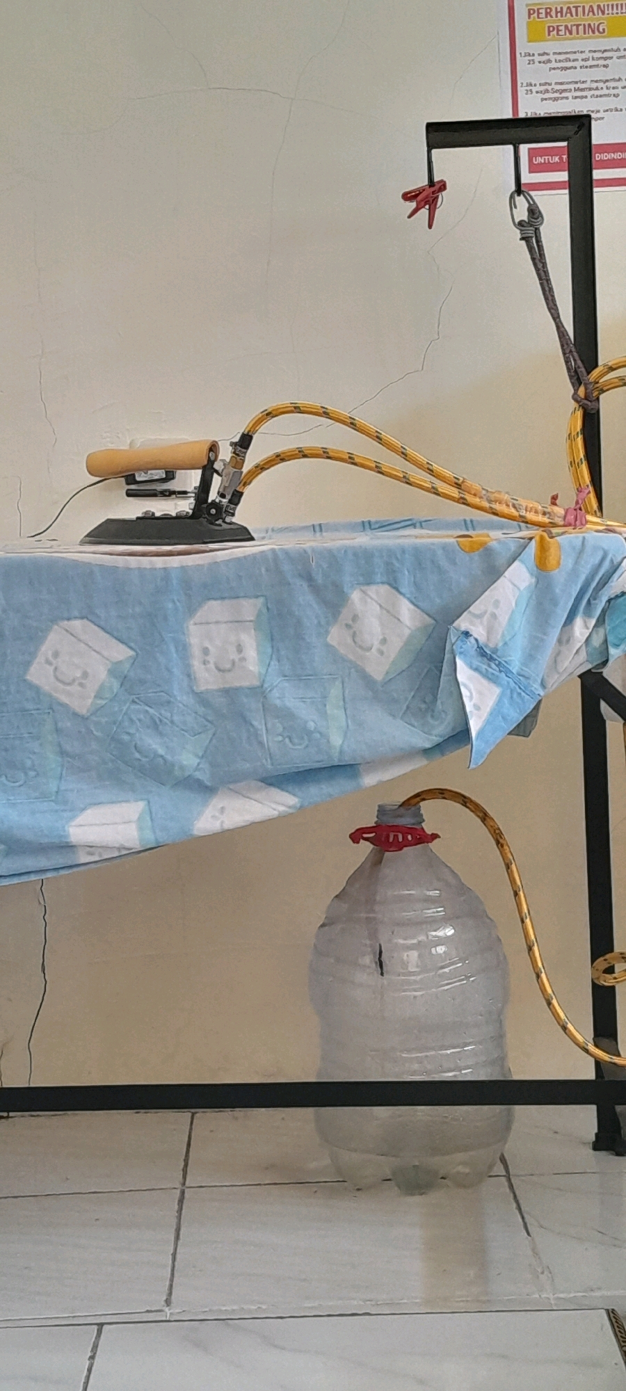 An indoor scene in Rejosari, Indonesia, shows a makeshift steaming setup. An electric iron with a wooden handle rests on an ironing board covered with a blue and white sheet featuring cartoon cube designs. Two yellow hoses extend from the iron, one likely for steam and the other for water. These hoses are attached to a black metal stand that also supports a red clothespin. Another yellow hose snakes down to a large, clear plastic bottle filled with water, capped with a red plastic fitting. The bottle sits on a tiled floor. A poster with Indonesian text is partially visible on the wall in the background, featuring the word "PENTING" (Important). The wall itself appears to have hairline cracks.