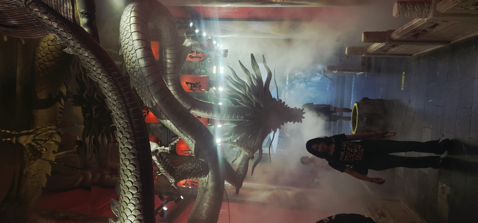 A woman in a black t-shirt and dark pants is walking towards the camera in a dimly lit indoor space. She is smiling and has a black purse slung over her left shoulder. In front of her is a stone basin. Behind her, a large, dark dragon sculpture snakes through the space, with red lanterns hanging from the ceiling. Smoke or fog fills the air, obscuring some of the background details and other people who are further away. The floor is made of dark stone tiles. The overall atmosphere suggests an entertainment venue or themed attraction, possibly in Yuzhong District, China, given the dragon motif and red lanterns often associated with Chinese culture.