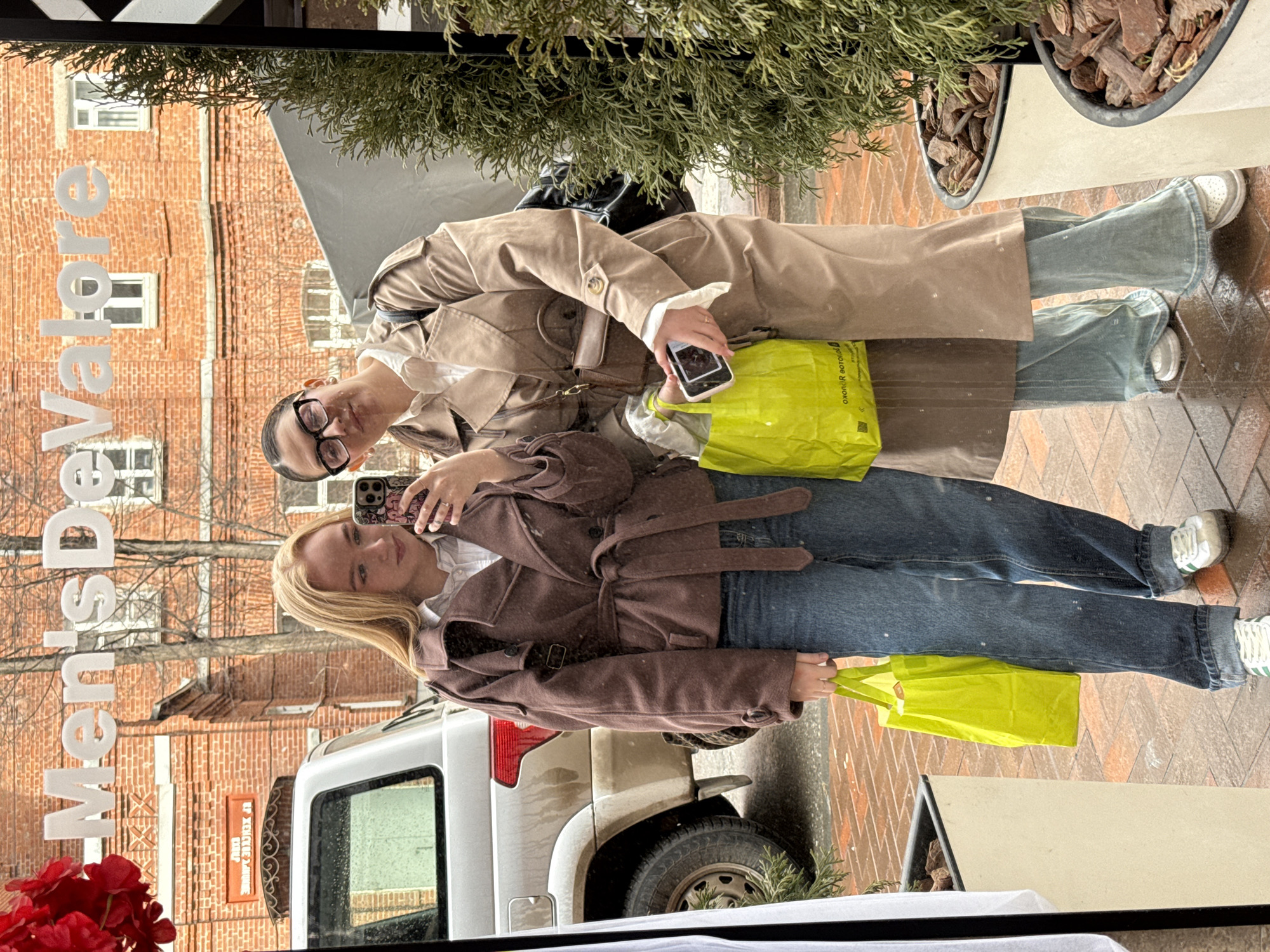 Two young women are reflected in a shop window in Perm, Russia. One woman, on the left, has blonde hair and is wearing a brown jacket, jeans, and is holding a yellow shopping bag. She is taking a selfie. The other woman, on the right, has dark hair, is wearing glasses, a tan trench coat, jeans, and is holding a cell phone and a yellow shopping bag. A light-colored car is partially visible. Behind them, the reflection shows a brick building with windows and the word "MensDevatore" written backward. In the upper part of the reflection, greenery is visible, likely from a plant or tree. The bottom of the image shows a black border and part of a white surface. The environment appears to be a city street.
