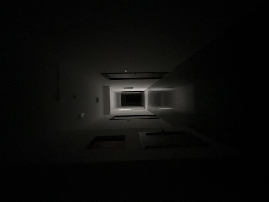 The image depicts a long, dim hallway located in Boreti, Montenegro, viewed from a slightly elevated, head-on perspective. The foreground and immediate sides of the corridor are enveloped in deep shadow, with light gradually increasing towards the distant end, creating a strong one-point perspective. The floor is light-colored and reflective, casting faint vertical reflections of the distant light source. Light-colored walls are discernible in the middle ground, contrasting with several dark-colored doors lining both sides of the hallway; at least three are visible on the left and two on the right. On the left wall, closer to the foreground, a framed rectangular object and a lower access panel or door are partially visible. The ceiling in the illuminated section features several circular fixtures, possibly smoke detectors or dormant lights. The scene is devoid of individuals, active movement, or discernible interactions.