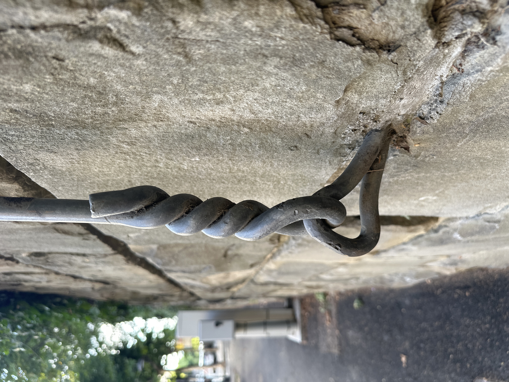 This is a close-up, eye-level, exterior, daylight shot of a weathered, dark grey metal chain link that is attached to a rough, beige stone wall. The chain is twisted and appears to be made of thick, solid metal rods. The stone wall is constructed of large, irregular blocks, showing signs of age and wear. To the left of the wall, a paved pathway is visible, with a faint suggestion of green foliage and trees in the background. The overall scene is devoid of people. The perspective suggests the chain is a functional element, possibly for a barrier or support, embedded into the wall. The location is identified as being in the city of Trieste, Italy.