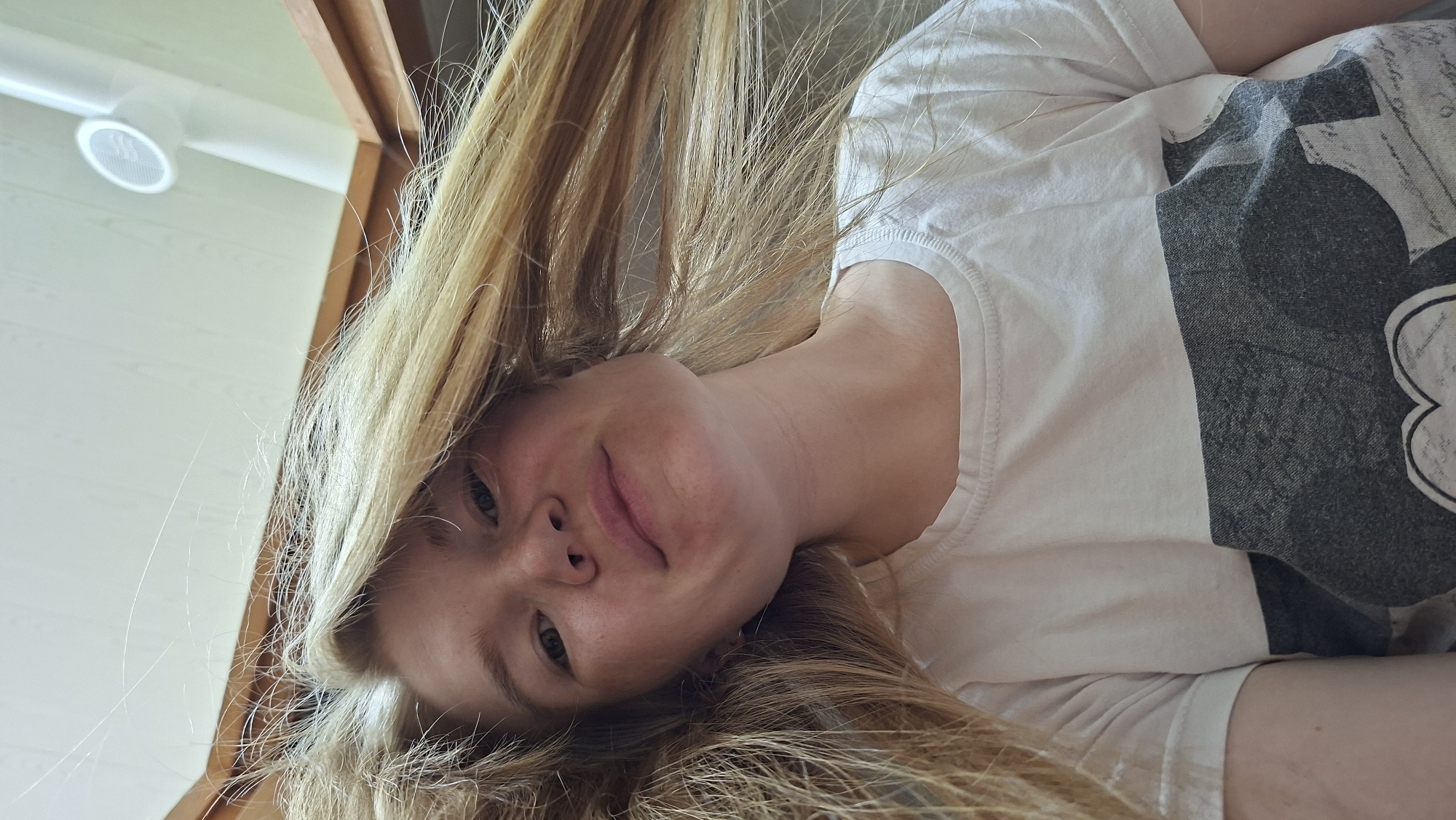 The image is a low-angle, close-up selfie of a young woman with blonde hair, taken indoors. She is wearing a white t-shirt with a dark graphic print on the left side. The lighting suggests an indoor environment, possibly a room with a ceiling fan visible in the background on the right. The woman's face is tilted downwards, and she is looking towards the camera with a slight smile. Her hair is long and falls around her face and shoulders. The background consists of wooden beams and a white, wood-paneled ceiling. The image does not contain any elements that would allow for the identification of the specific location in the city of Большой Букор, Russia.
