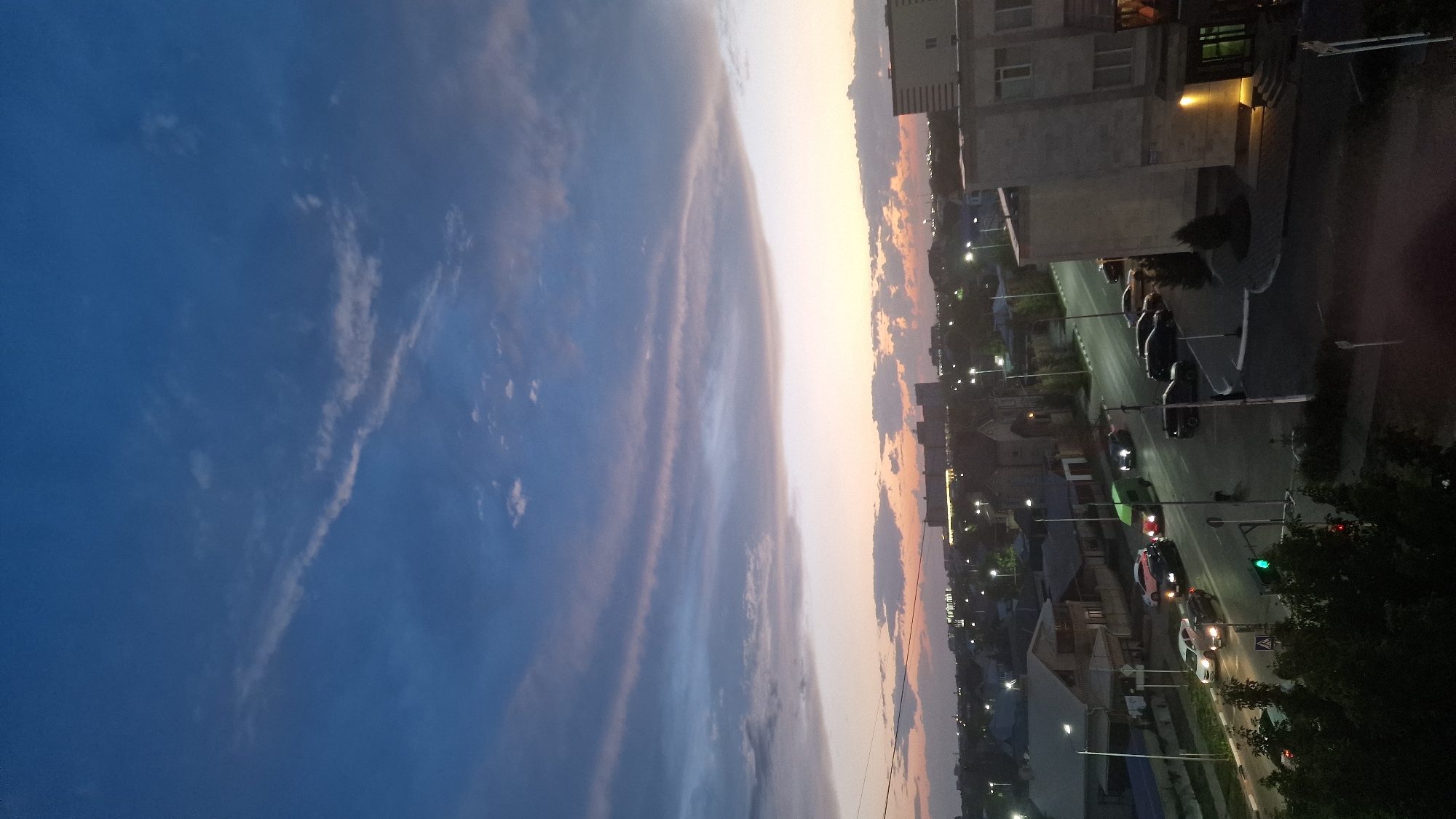 The image captures a twilight scene in Kostanay, Kazakhstan, overlooking a residential and commercial area. The sky is a gradient of deep blue, transitioning to pink and orange hues near the horizon, with distinct layers of clouds. Below, a city skyline is visible, characterized by various buildings, including what appears to be a large, modern structure on the right and smaller, more traditional residential houses with pitched roofs. Streetlights are illuminated, casting pools of light along the roads and sidewalks. A multi-lane road runs through the scene, with several cars in motion, some appearing to be at an intersection with traffic lights. The foreground shows some greenery, suggesting the presence of trees or bushes. The overall atmosphere is calm, indicative of the early evening hours after sunset.
