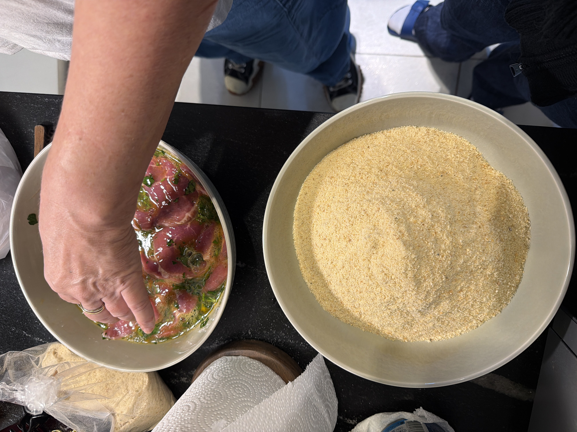 An overhead shot depicts a person's arm reaching into a bowl of sliced meat mixed with marinade. Adjacent to it is a bowl filled with a tan granular substance, likely breadcrumbs. The setting appears to be indoors, possibly a kitchen, based on the countertop surface and paper towels nearby. The presence of legs wearing jeans and shoes suggests additional individuals are present in the scene. The location is Vicente López, Argentina.