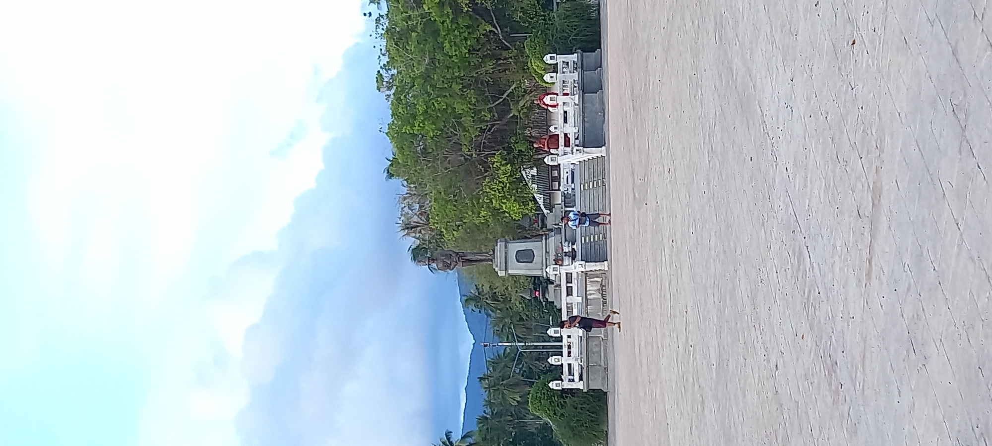 Captured from an elevated viewpoint, the photograph shows a sunlit scene in Thong Sala, Thailand, featuring a statue of a soldier standing on a pedestal. The statue faces toward lush green trees and a distant mountain range beneath a sky filled with white clouds. A series of white fences and stairs lead up to the statue. Several individuals, some dressed in red, are observed standing or walking near the stairs. The foreground shows a concrete pavement with diagonal scoring marks.