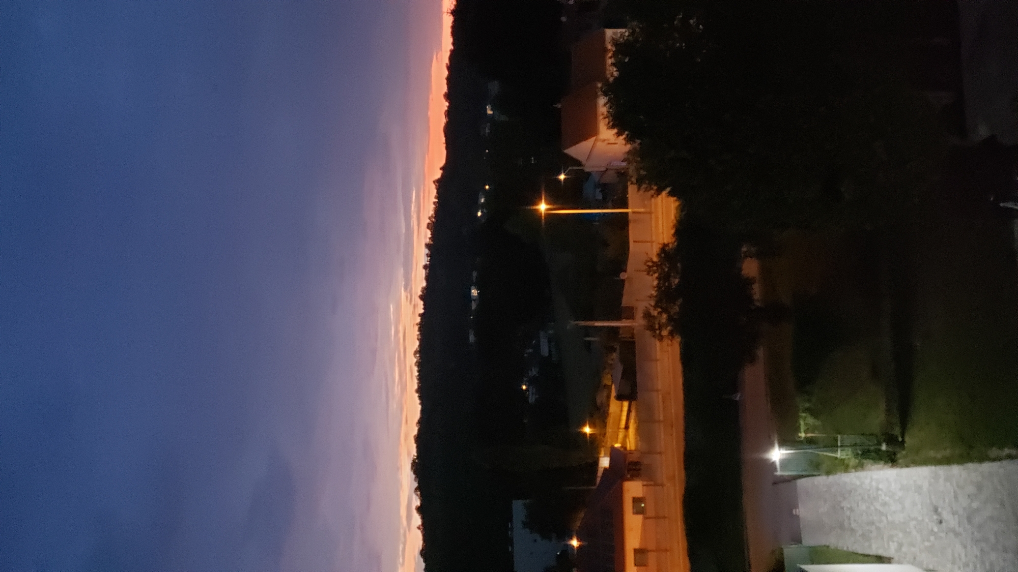 The image depicts a view of a town at dusk, likely in Prešov, Slovakia. The sky is a deep blue transitioning to purple and orange hues near the horizon. Silhouetted against the twilight are rolling hills covered in trees. In the foreground and midground, there are several residential buildings and streetlights illuminated. One building in the mid-left has a brown roof and lights on inside. A road runs through the scene, bordered by a fence and illuminated by streetlights. Further back, more houses and lights are visible scattered amongst the trees. The foreground shows a cobblestone path leading away from the viewer, with a lamppost nearby. The overall impression is a quiet, residential area settling in for the evening.