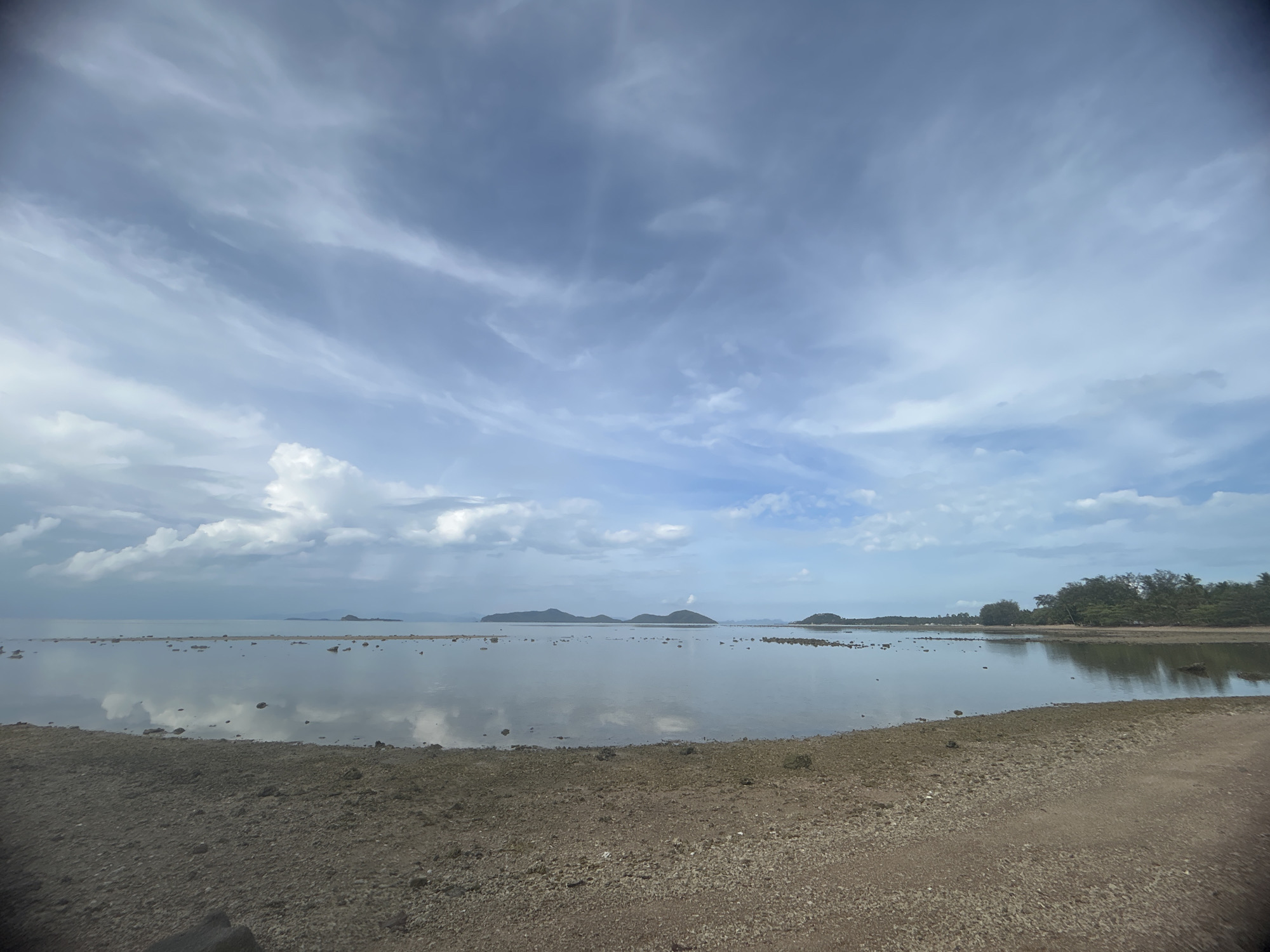 The image captures a tranquil seascape in Ban Bang Khao, Thailand. The foreground features a beach comprised of gravel and sand. Calm, shallow water extends from the beach to the horizon, reflecting the sky above. Several small islands are visible in the distance, appearing as dark silhouettes against the lighter sky and water. On the right side of the frame, trees line the coast. Above, the sky is filled with a mix of clouds, with patches of blue visible in between. The overall lighting suggests an overcast day.