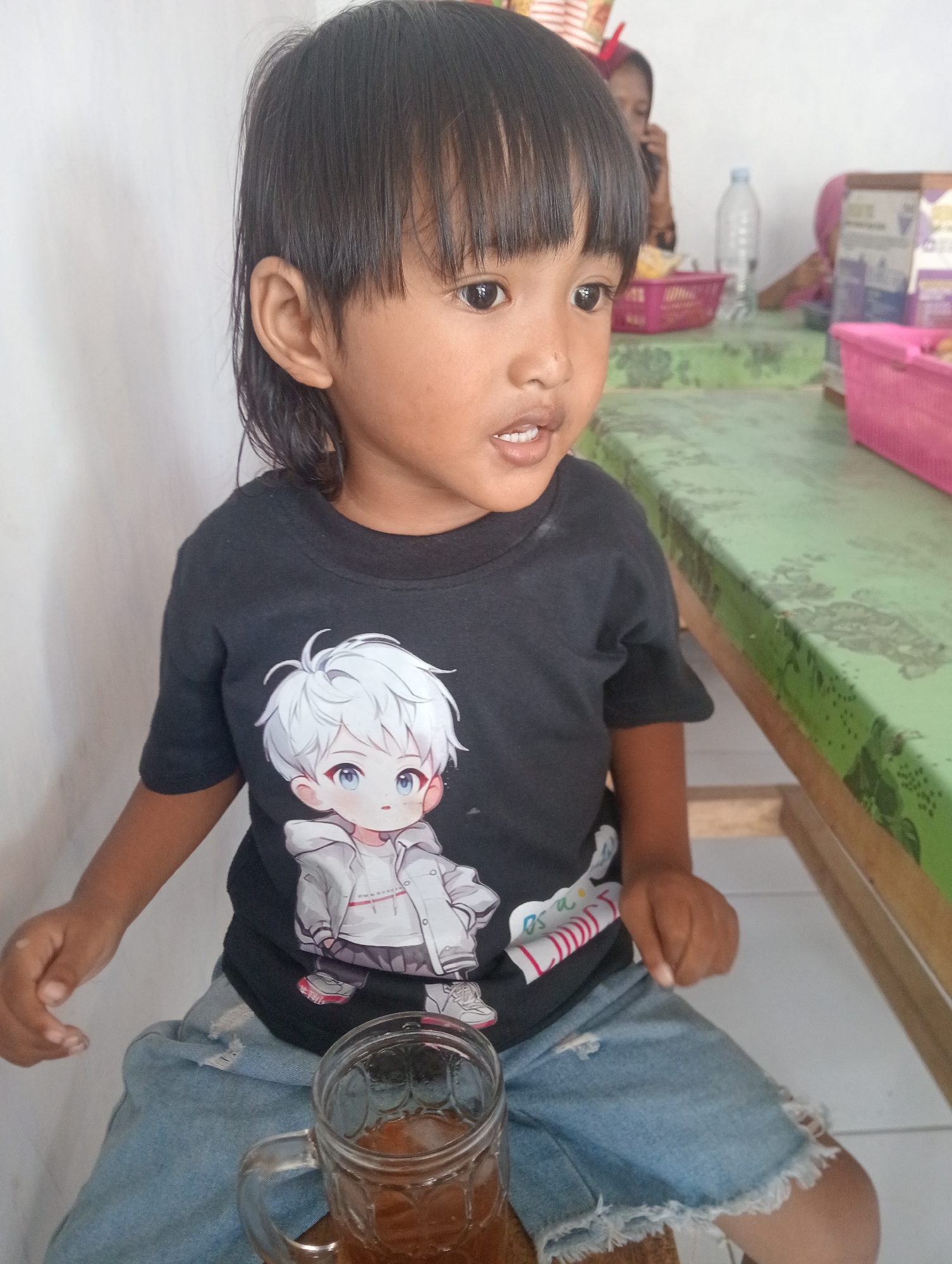 A young girl with black hair and bangs sits at a table, wearing a black t-shirt with a cartoon image of a white-haired boy and blue denim shorts with ripped hems. She holds a glass mug filled with a brown beverage and ice. In the background, two other individuals, partially visible, are seated at the table, and pink plastic baskets are present. The environment suggests a casual indoor setting, possibly a restaurant or home, in Besuki, Indonesia.