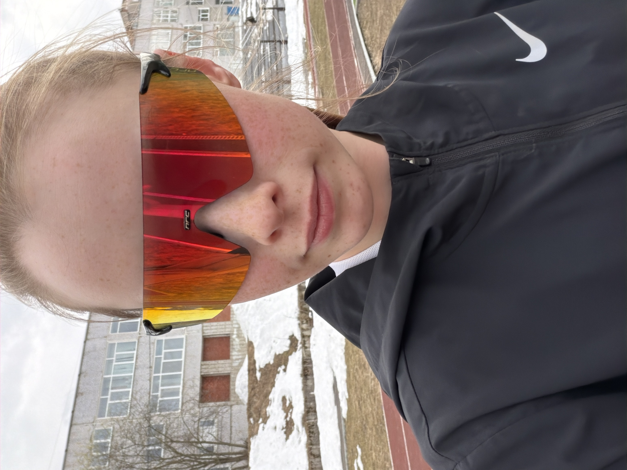 A slightly rotated outdoor portrait shows a young person with light hair and freckles, wearing large, mirrored sunglasses with red, orange, and yellow gradient lenses. Their head is tilted to the right, and their lips are slightly parted. The sunglasses obscure their eyes. They are wearing a dark grey or black zip-up jacket with a white Nike swoosh logo visible on the left side of the chest. A white collar or undershirt is visible underneath. A small logo, possibly "nfc", is visible on the lower left frame of the sunglasses lens.
The background features a large, multi-story building with numerous windows, some appearing discolored or covered. The building's architecture suggests an older, possibly Soviet-era, concrete structure. In the foreground and middle ground, patches of snow are visible on the ground, indicating cold weather. A reddish-brown running track is also visible, suggesting a sports facility or schoolyard. Bare tree branches are scattered throughout the background. The sky appears overcast, and the overall scene suggests daytime in a cold climate, consistent with the location context of Berezniki, Russia.