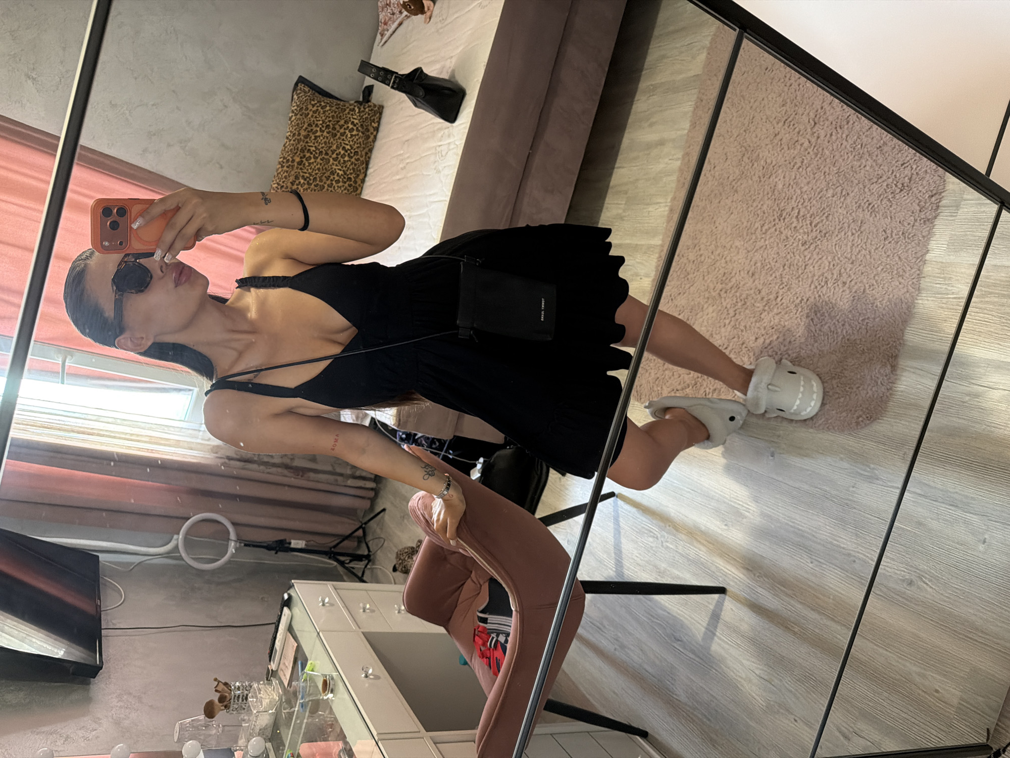 A woman is taking a mirror selfie in a bedroom. She is wearing a black dress, white shark slippers, and sunglasses. She is holding an orange phone in her right hand. In the background, there is a bed with a leopard print pillow, a vanity with makeup, and a pink chair. The room appears to be a residential bedroom. The location is not specified in the image.
