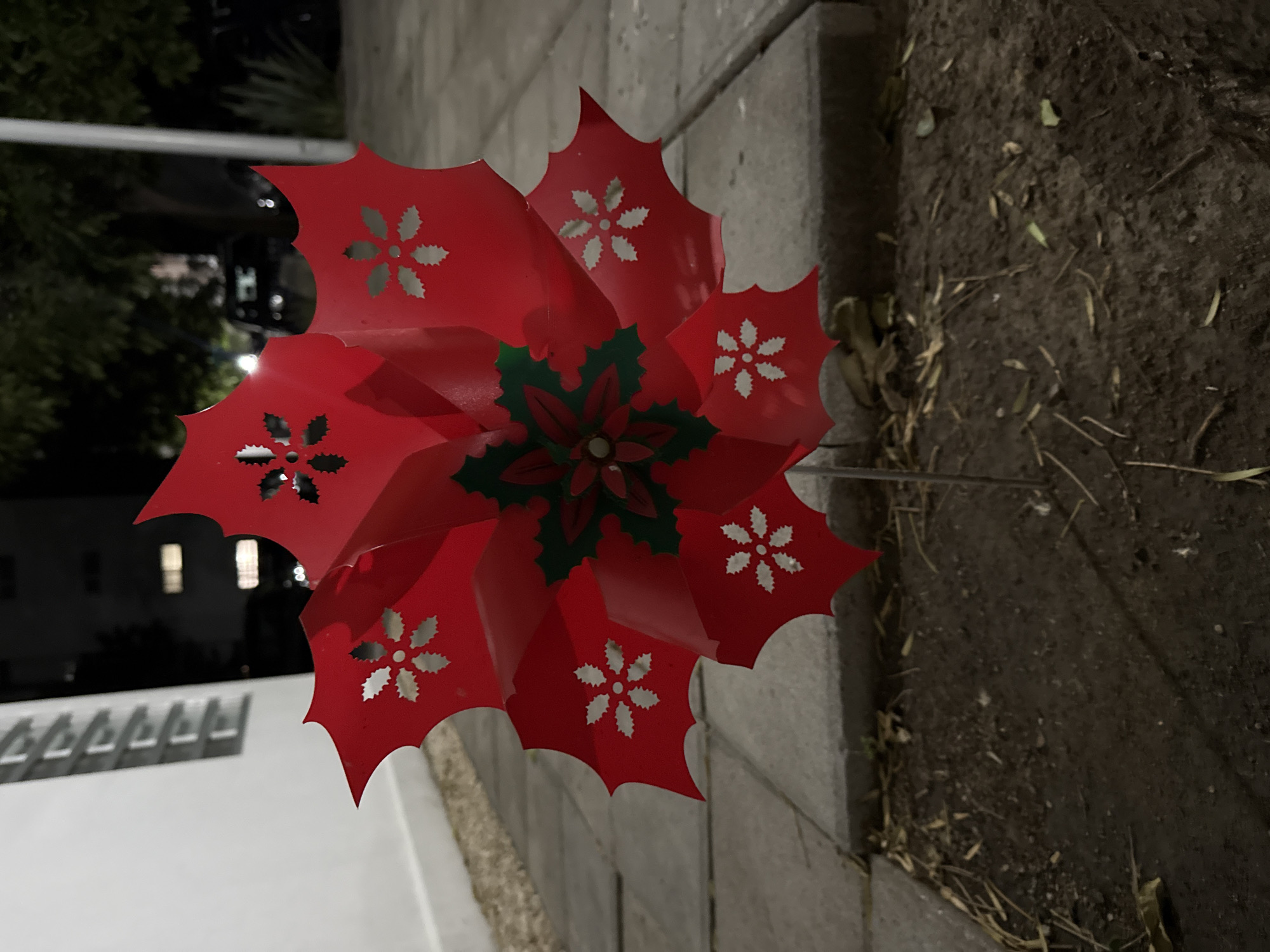 A red poinsettia pinwheel stands on a metal rod in a patch of dirt adjacent to a cement walkway. The pinwheel has cut-out flower designs on each of its petals. The central part of the poinsettia is green. The scene is at night. A white building with square details is behind the pinwheel. The exact location in the city of Puebla, Mexico is impossible to determine without further information.
