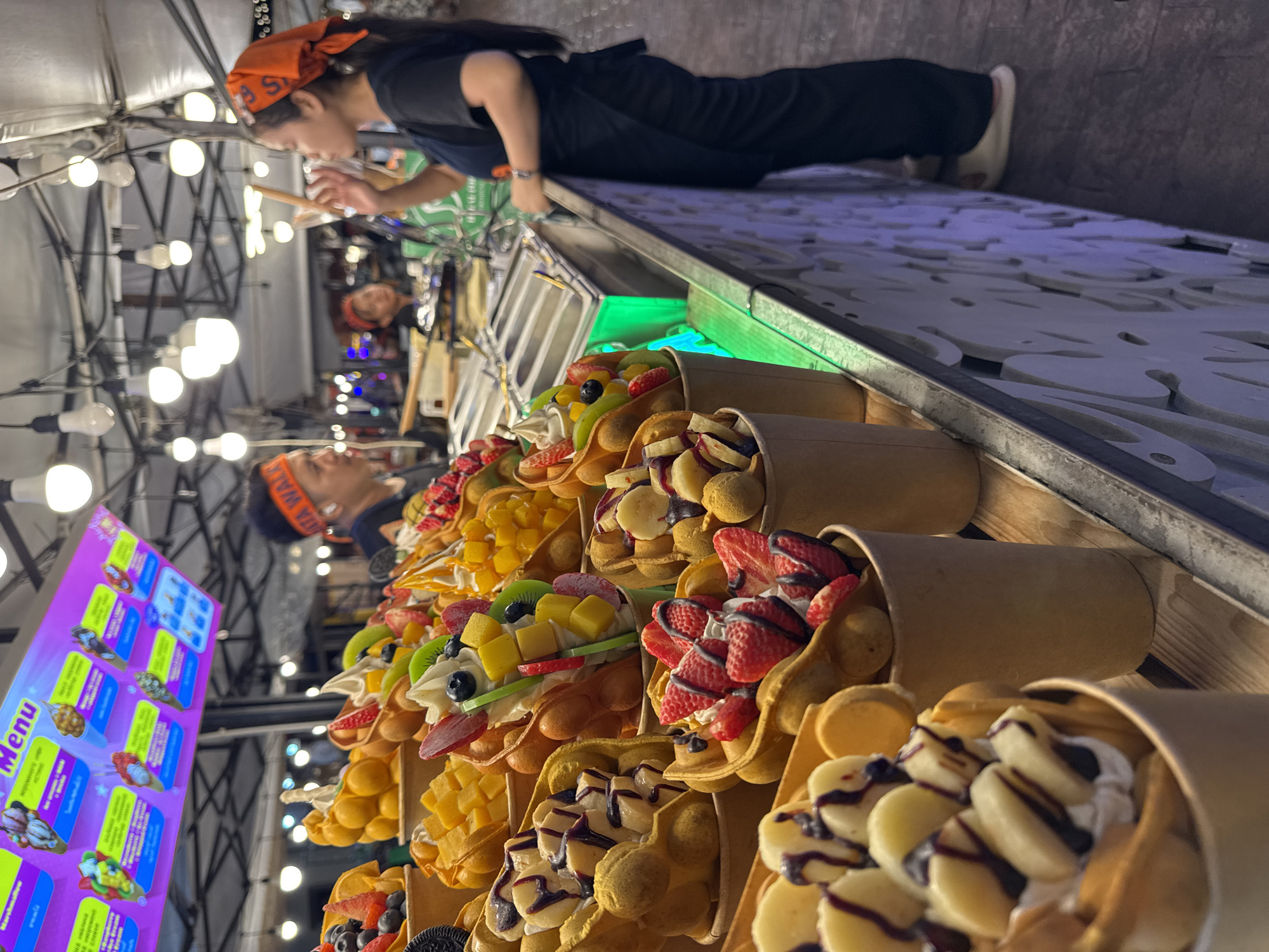 The image shows a food stall in Karon, Thailand, with a variety of bubble waffles with fruit toppings displayed. A woman wearing an orange bandana and black attire is seen preparing or serving the waffles. The stall is brightly lit, possibly at night or in a shaded area, with numerous light bulbs visible. A menu board with colorful images of the waffles is displayed on the left side of the image. The waffles appear to be filled with a creamy base and adorned with fresh fruits such as strawberries, blueberries, kiwi, mango, and bananas, along with chocolate syrup. The setting suggests a bustling street food market or a dessert shop. The overall mood appears lively and appetizing. The menu board has text in what appears to be a Southeast Asian language, with "Menu" clearly visible in English.