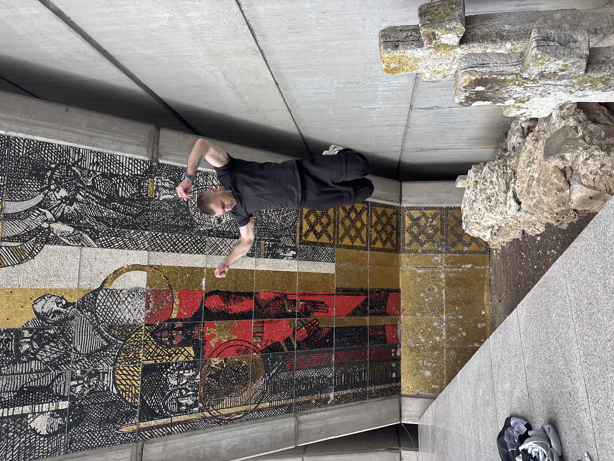The image displays a man lying on his back on a light grey concrete floor, positioned in front of a large, colorful mosaic wall. The man, wearing a black t-shirt, black pants, and white sneakers, has his left arm bent over his head and his right arm extended towards the mosaic. His gaze is directed upwards or away from the camera.
The mosaic, which covers a significant portion of the wall, is composed of numerous small tiles in shades of black, white, gold, and red. It features intricate, detailed patterns that include both abstract geometric designs and figurative elements, some resembling faces or historical scenes. Above the mosaic, there is a concrete beam and a plain concrete wall or ceiling.
To the right of the man and further into the background, several large, weathered stone structures are visible. These structures appear to be ancient or stylized, with some resembling crosses or carved pillars, displaying natural discoloration or moss. Below them are more rugged, natural rock formations. In the foreground, on the concrete floor near the bottom right, a pile of folded clothes or fabric items can be seen. The overall setting suggests a sheltered outdoor area, such as an underpass, a public monument, or a building's exterior, characterized by a blend of modern concrete architecture, historical-looking mosaic art, and natural stone elements. The lighting is soft and even, implying an overcast day or a well-lit indoor environment. No discernible text is visible.
