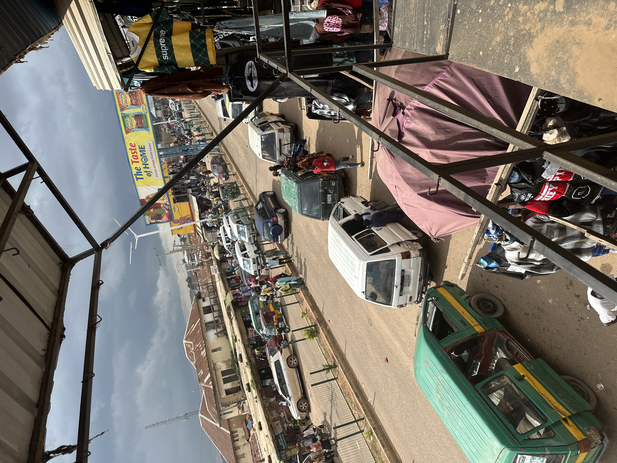 This image depicts a busy street scene in Bukuru, Nigeria, under a cloudy sky. The foreground features a stall with clothing items hanging, including shirts and jackets. A dark purple tarp covers a structure adjacent to the stall. In the middle ground, a green van with yellow stripes is parked on the street, its doors open. Several other vehicles, including white vans and cars, are also present on the road, which is lined with shops and buildings. A large billboard with the text "The Taste of HOME" and product advertisements is visible in the background, along with a radio tower. Pedestrians are seen walking along the street and sidewalks, and a few people are interacting with vendors. The overall environment suggests a bustling marketplace or commercial area.
