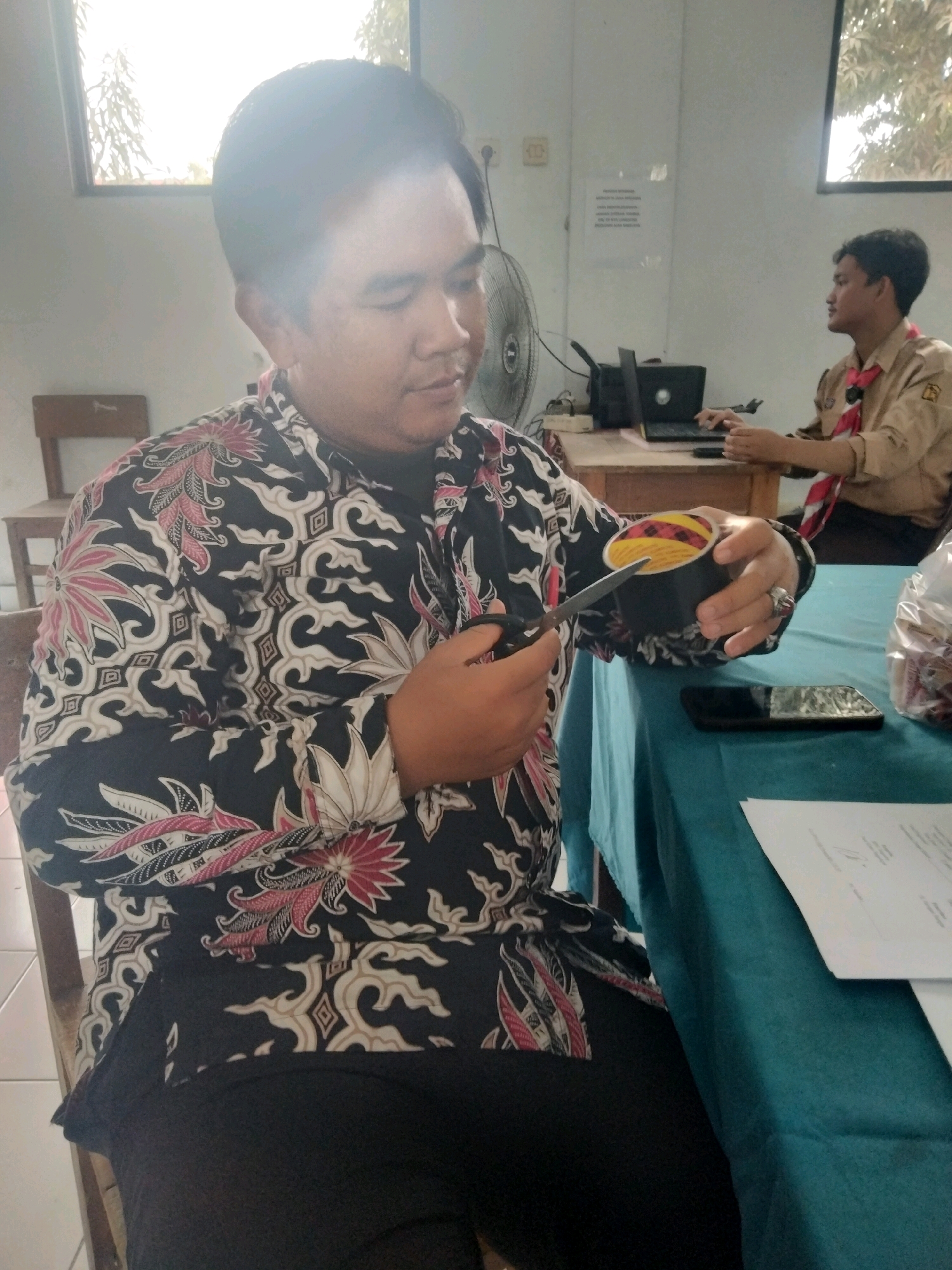 In the city of Suka Sari, Indonesia, a man in a black batik shirt with pink and white floral patterns is sitting at a table. He is holding a pair of scissors in his right hand and appears to be cutting a cylindrical object that contains colorful striped material. His left hand is supporting the object. He is wearing a ring on his left ring finger. A cell phone is lying on the teal tablecloth in front of him. In the background, another person wearing a tan uniform with a red scarf is seated at a desk with a laptop. The room appears to be an indoor space with a window showing trees outside.
